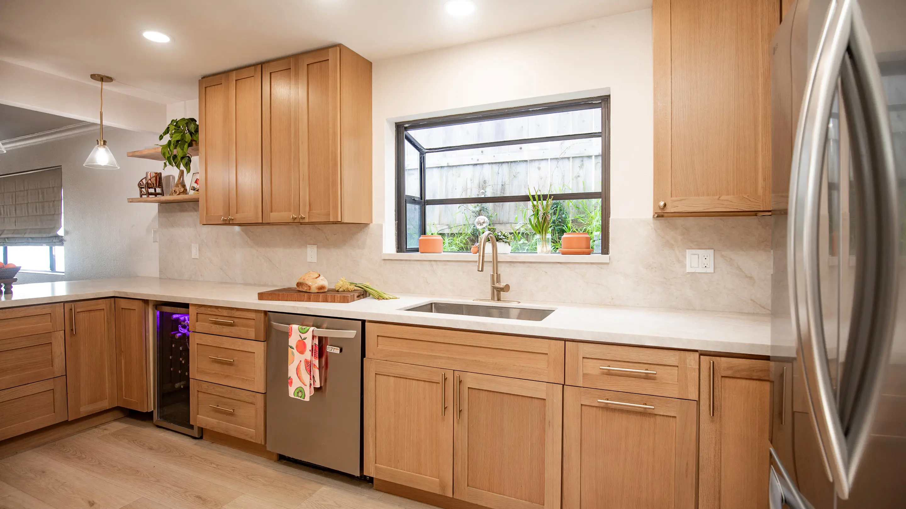 Kitchen by Sunshine Collective Studio with warm shaker cabinets, marble slab backsplash, stainless undermount sink, wine fridge, black-framed garden window, and floating wood shelf with trailing pothos on light hardwood floors.