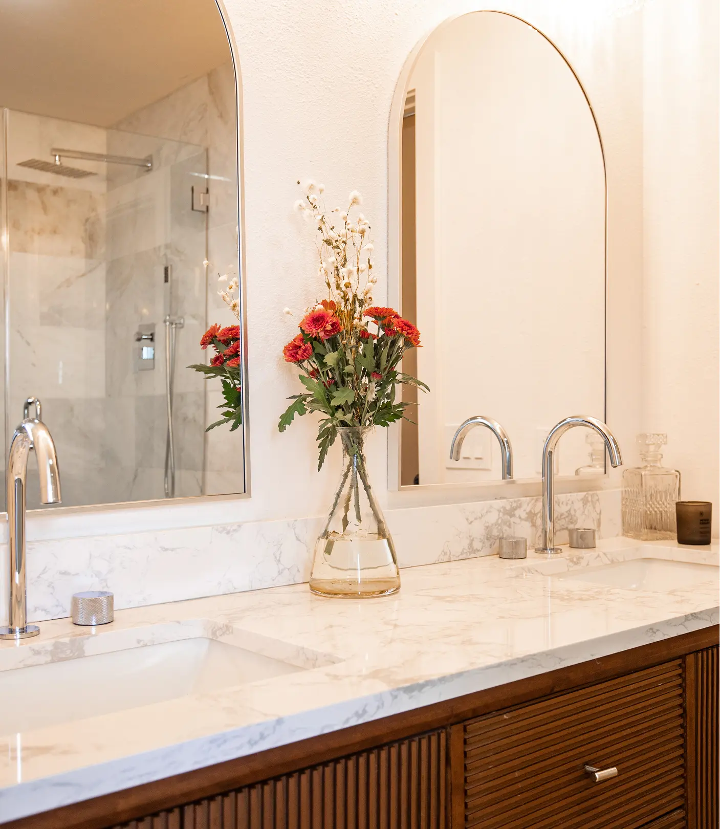 Primary bathroom vanity detail by Sunshine Collective Studio with dual arched mirrors, fluted walnut cabinetry, marble countertop, chrome gooseneck faucets, and a fresh floral arrangement with a marble shower reflected behind.