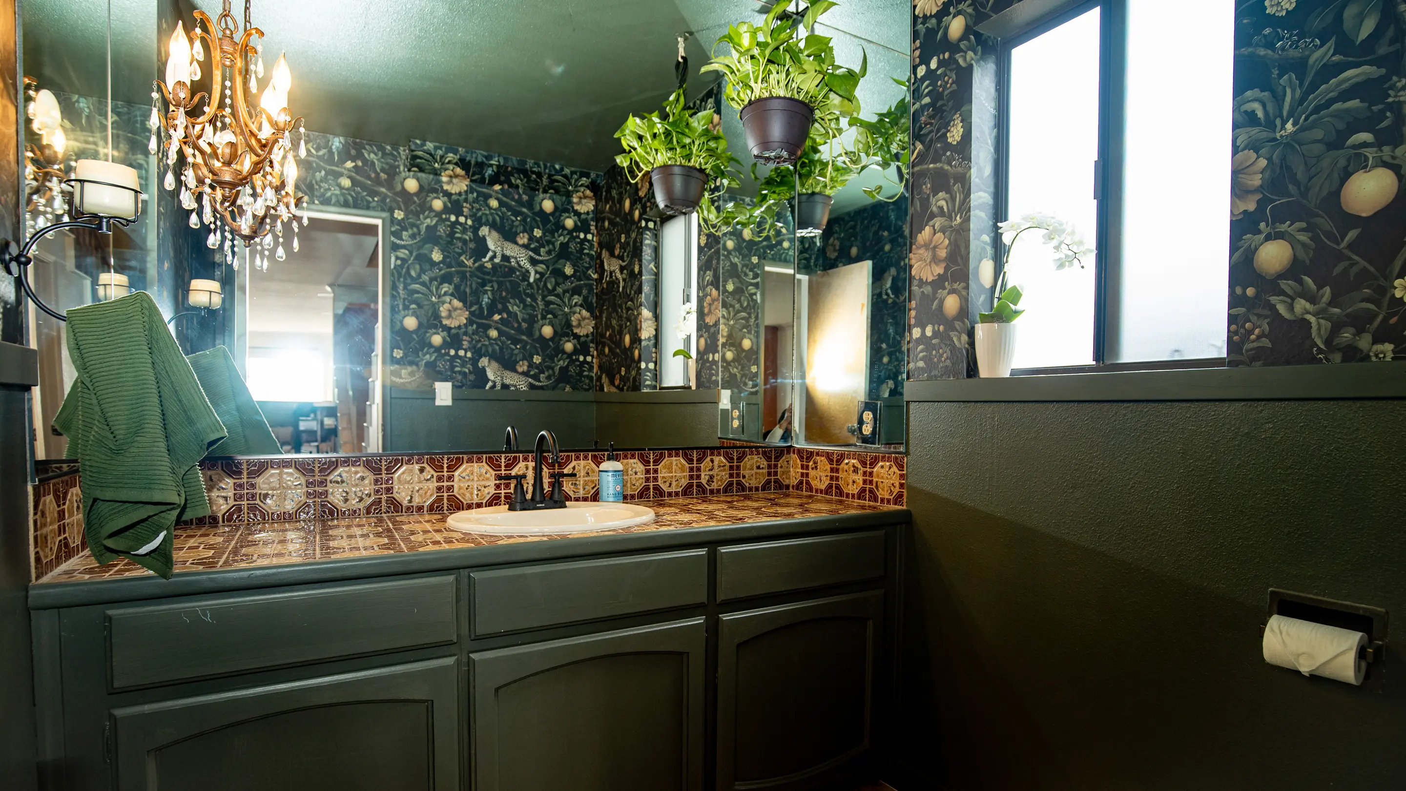 Moody powder room by Sunshine Collective Studio with dark botanical wallpaper, olive green cabinetry, decorative tile countertop, crystal chandelier, and hanging pothos plants above a black faucet sink.