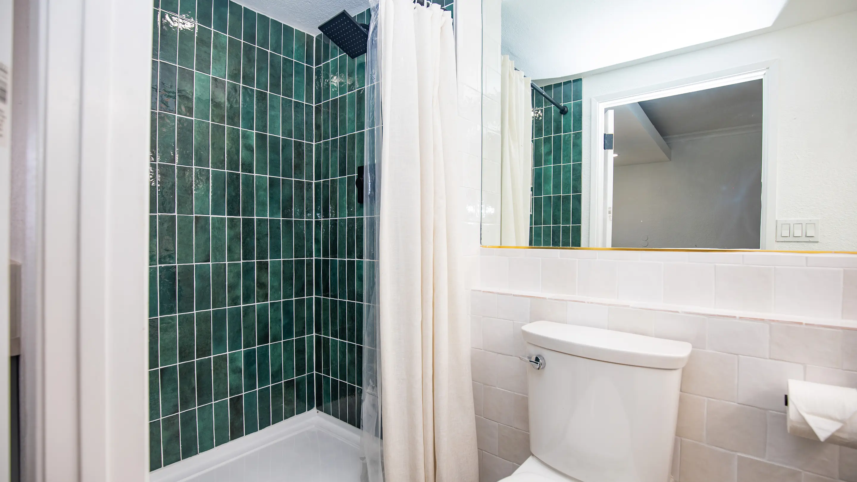 Bathroom by Sunshine Collective Studio featuring a bold deep green zellige tile shower with black rain head, white subway tile walls, linen curtain, and large mirror with gold trim accent.