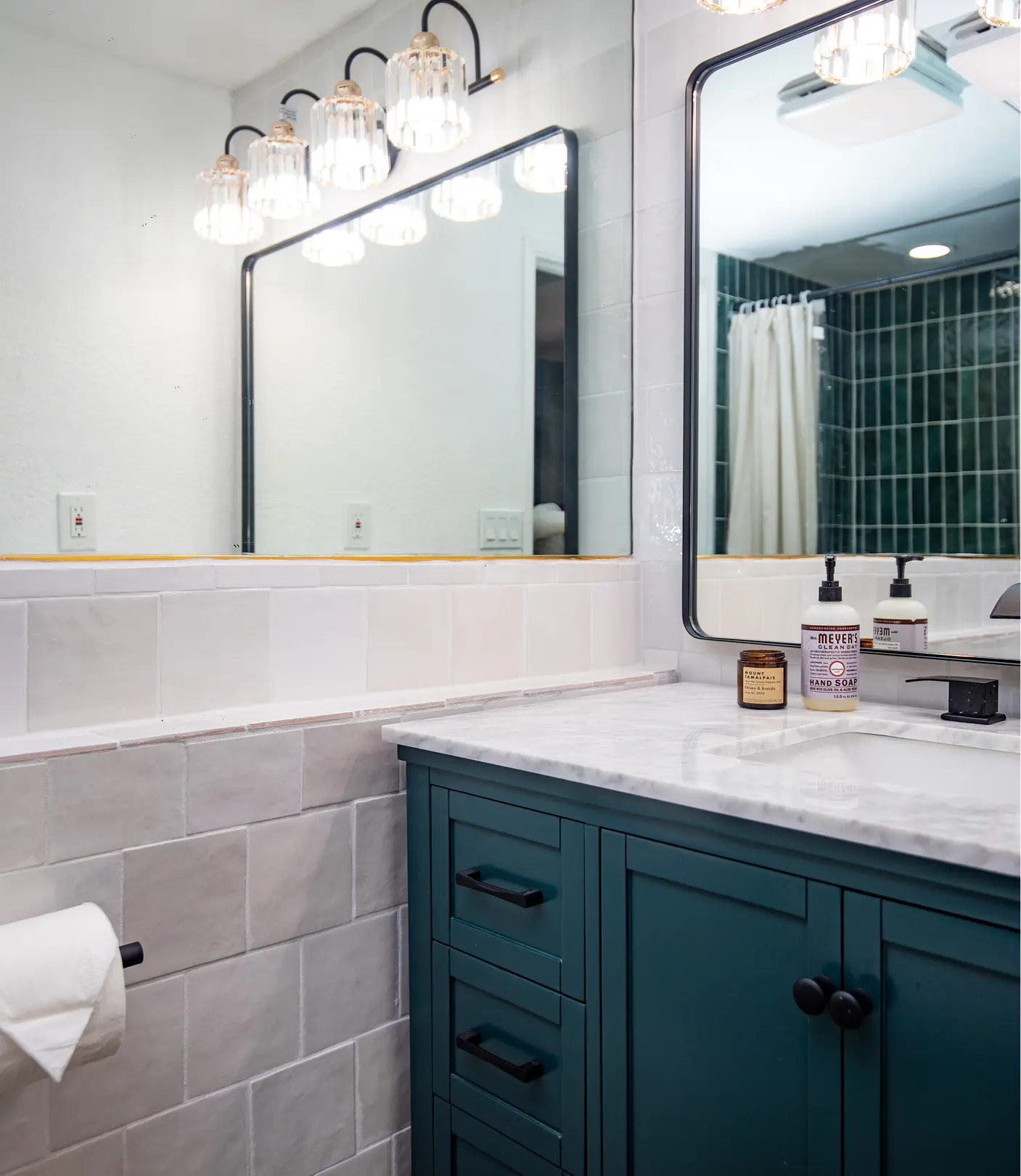 Renovated bathroom by Sunshine Collective Studio with a teal vanity, marble countertop, black-framed mirror, crystal globe sconces, white zellige tile walls, and deep green tile shower reflected in mirror.