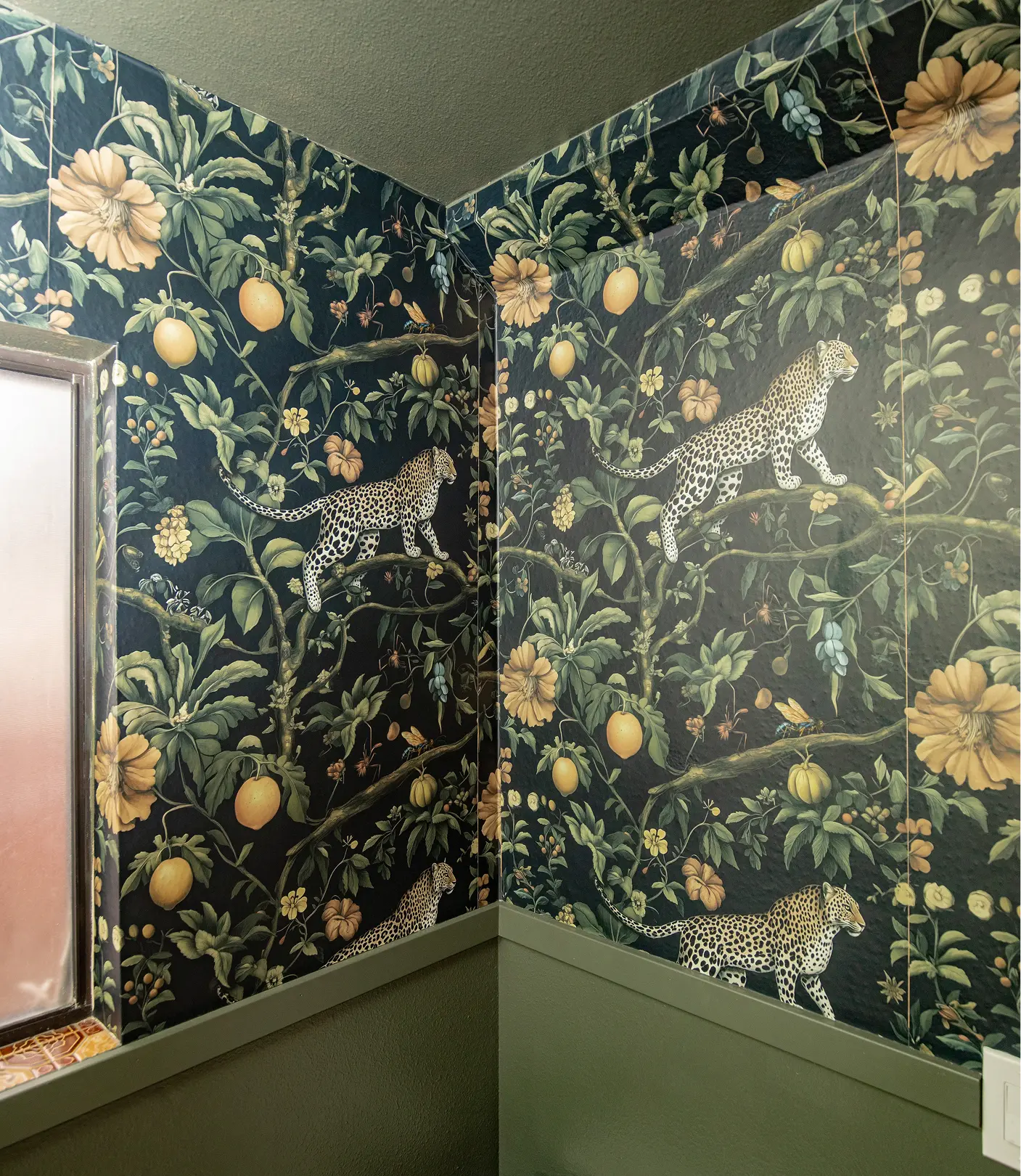 Wallpaper detail by Sunshine Collective Studio featuring a dramatic dark botanical print with leopards, citrus fruit, and oversized florals wrapping a corner above sage green wainscoting.
