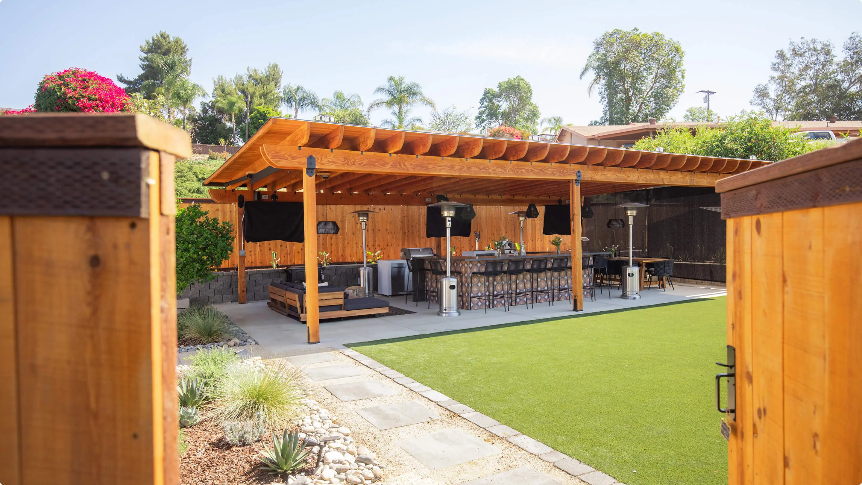 Backyard entertaining space by Sunshine Collective Studio featuring a large wood pergola with outdoor TVs, patio heaters, a tiled outdoor kitchen island with bar seating, and lush artificial turf surrounded by drought-tolerant landscaping.