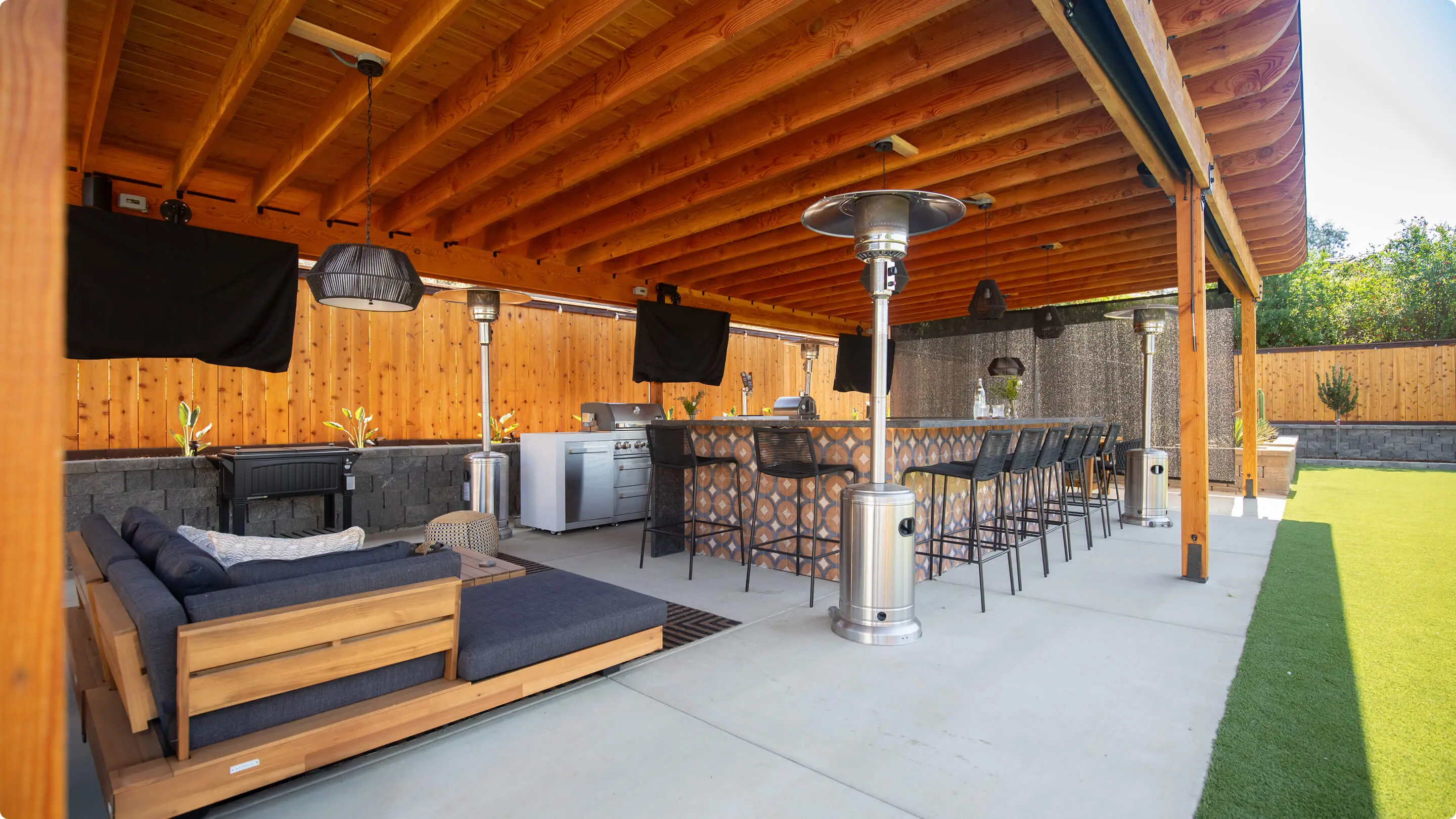 Covered patio by Sunshine Collective Studio with a wood pergola, teak sectional with navy cushions, tiled outdoor kitchen bar, black rope pendants, patio heaters, mounted TVs, and artificial turf.