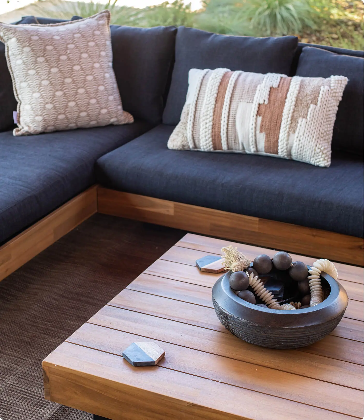 Outdoor lounge detail by Sunshine Collective Studio with a teak sectional and navy cushions, patterned throw pillows, a slatted teak coffee table, and a dark ceramic bowl with decorative orbs.