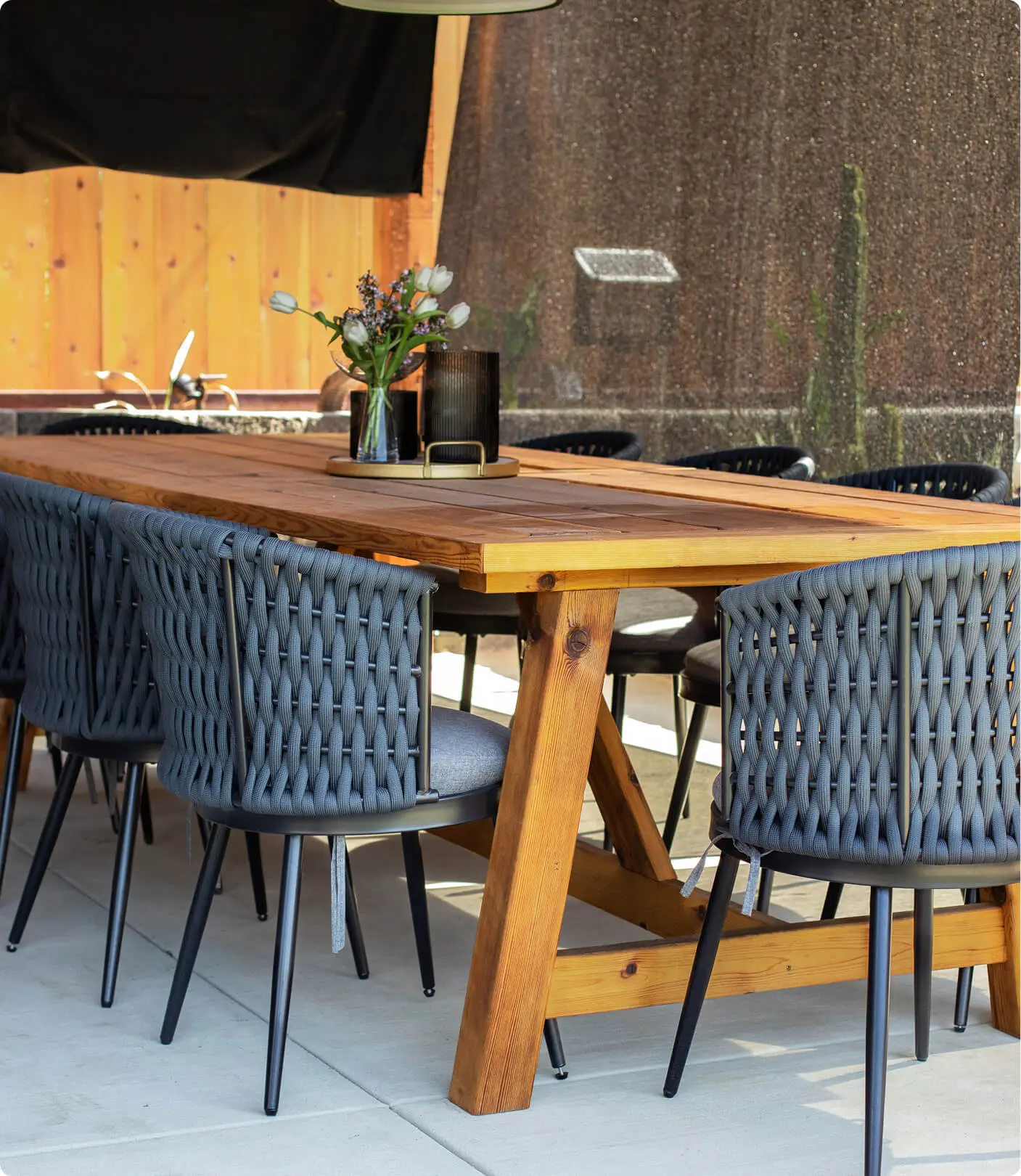Outdoor dining table detail by Sunshine Collective Studio featuring a solid wood trestle table with gray rope-back chairs and a gold tray centerpiece with white tulips and a smoked glass vase.