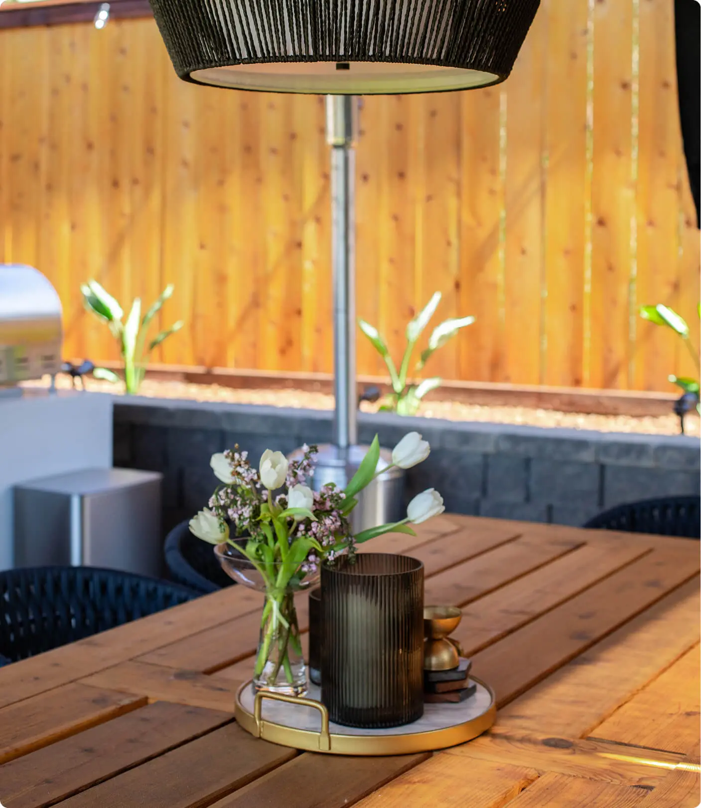 Outdoor table styling detail by Sunshine Collective Studio with a gold tray centerpiece holding white tulips, wildflowers, and a smoked ribbed glass, beside a black rope pendant light under a wood pergola.