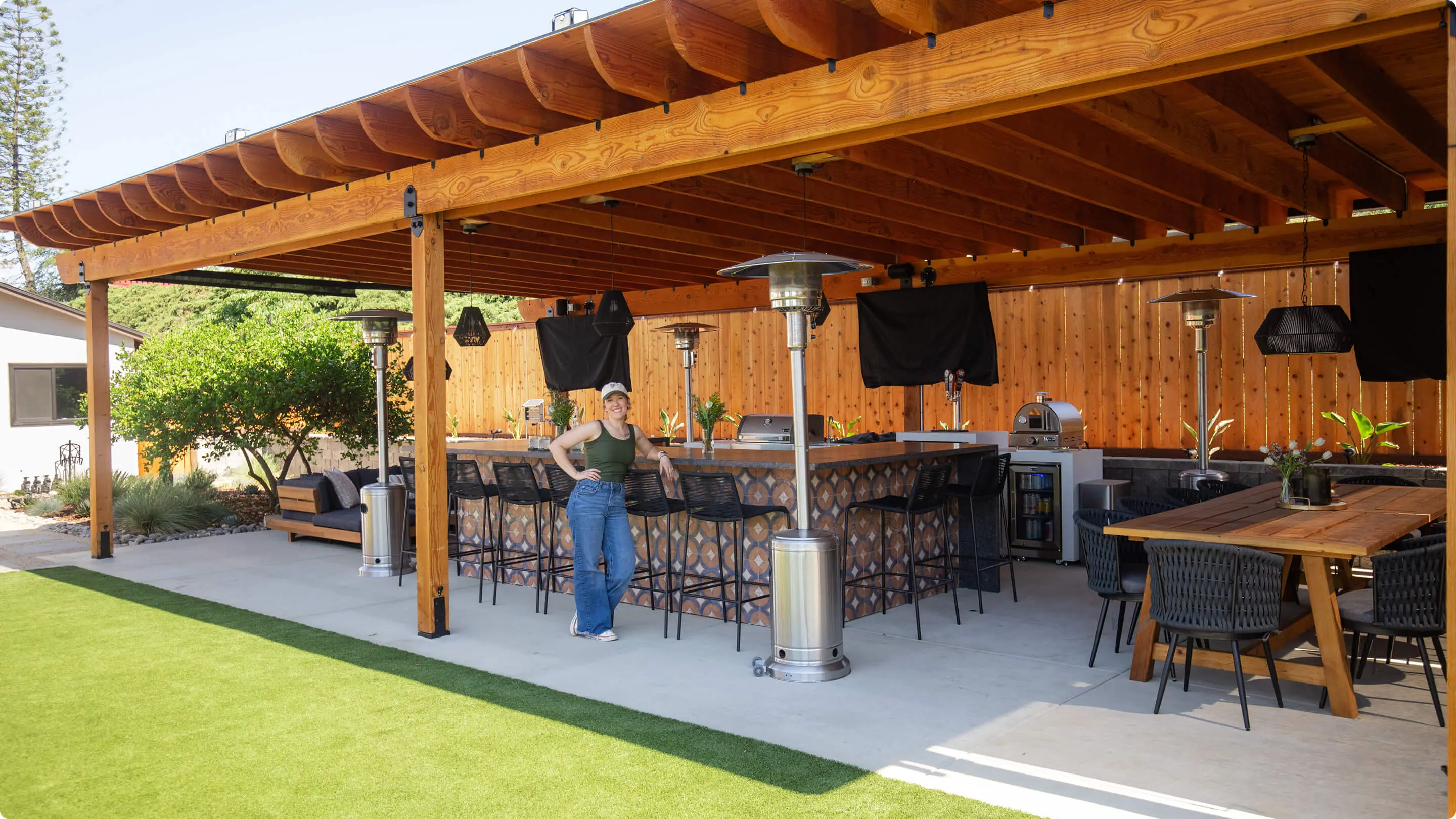 Founder and interior designer Kallie Morales of Sunshine Collective Studio stands beside a tiled outdoor bar island with pergola, patio heaters, mounted TVs, pizza oven, and teak dining table.