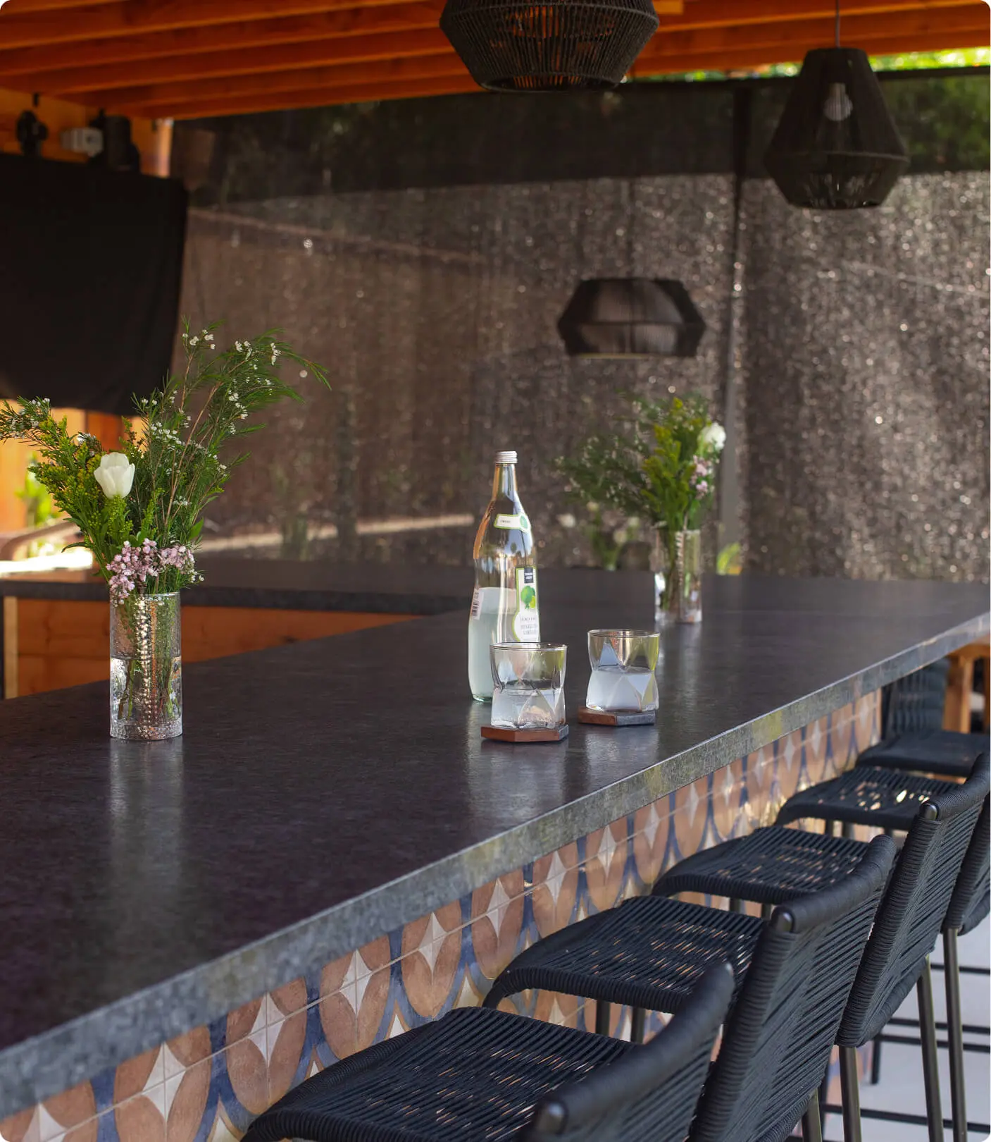 Outdoor bar detail by Sunshine Collective Studio featuring a dark stone countertop, decorative geometric tile fascia, black rope barstools, fresh florals, and a sparkling water bottle under black pendant lights.