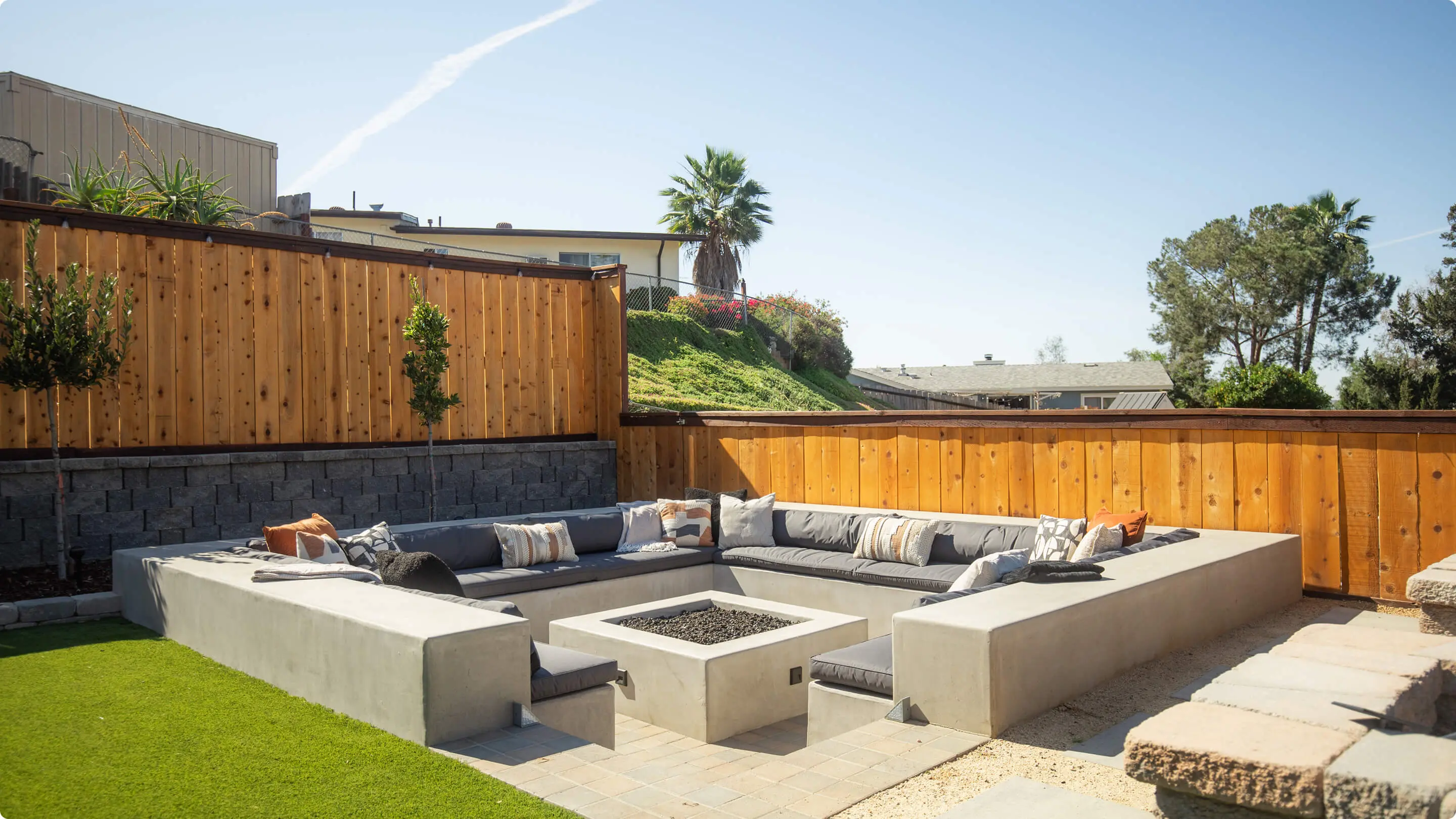Sunken outdoor lounge by Sunshine Collective Studio with a U-shaped concrete bench, gray cushions, patterned throw pillows, square concrete fire pit, cedar fence, topiary trees, and artificial turf.