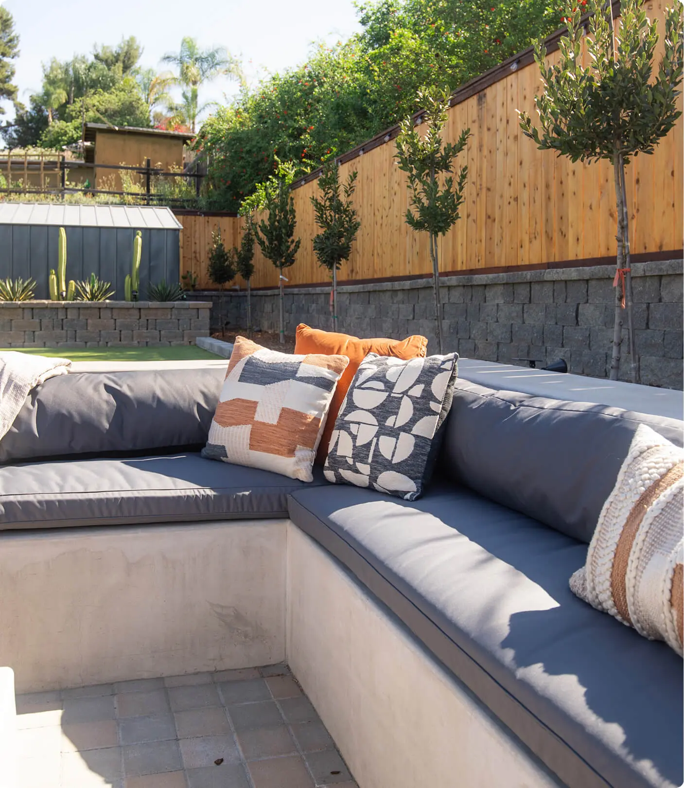 Built-in concrete bench detail by Sunshine Collective Studio with gray outdoor cushions and layered throw pillows in terracotta, geometric, and woven patterns, backed by topiary trees along a cedar fence.