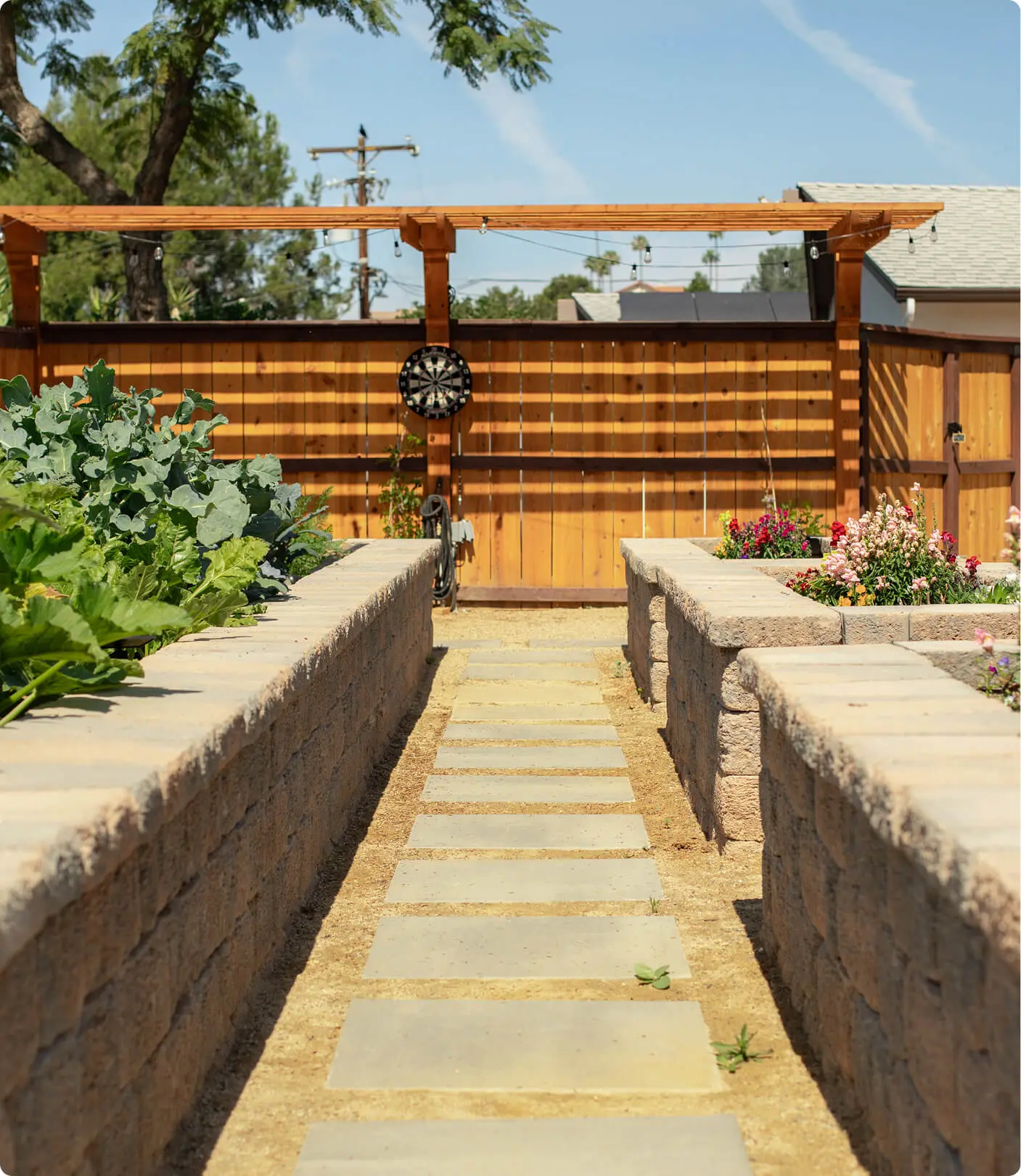 Garden pathway by Sunshine Collective Studio with a stone slab walkway between raised vegetable and flower beds, leading to a cedar pergola with string lights and a dartboard on the fence.