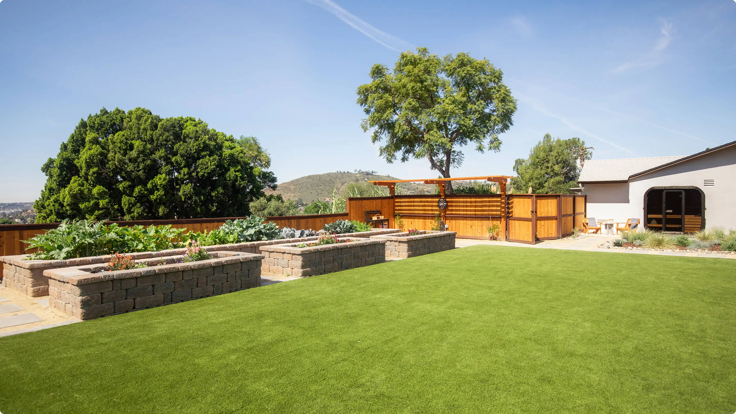 Backyard landscape by Sunshine Collective Studio with raised stone garden beds, lush artificial turf, a cedar pergola and privacy screen, an outdoor sauna, and sweeping hillside views under a clear blue sky.