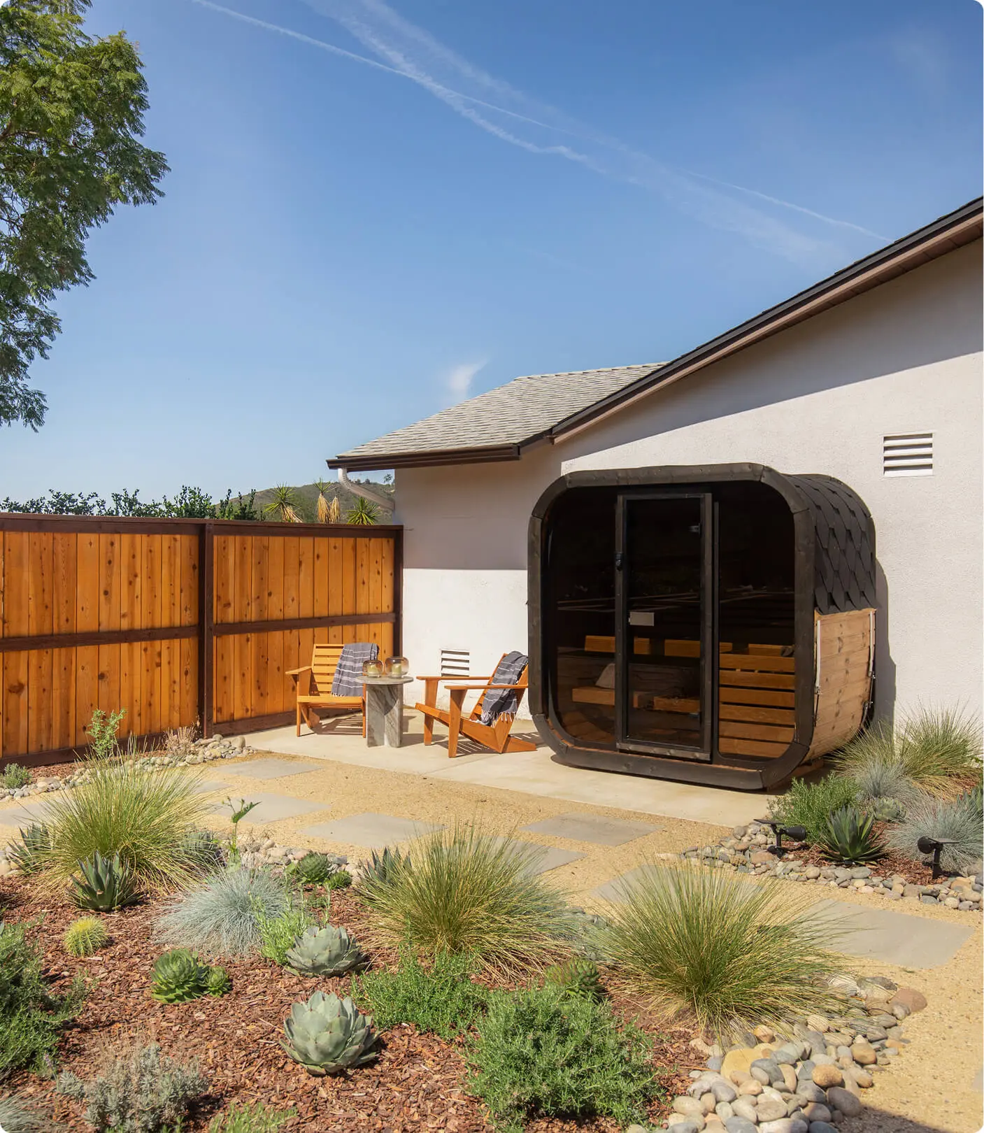 Outdoor sauna nook by Sunshine Collective Studio with a curved barrel sauna, two Adirondack chairs, a stone side table, and drought-tolerant landscaping with succulents, grasses, and agave.