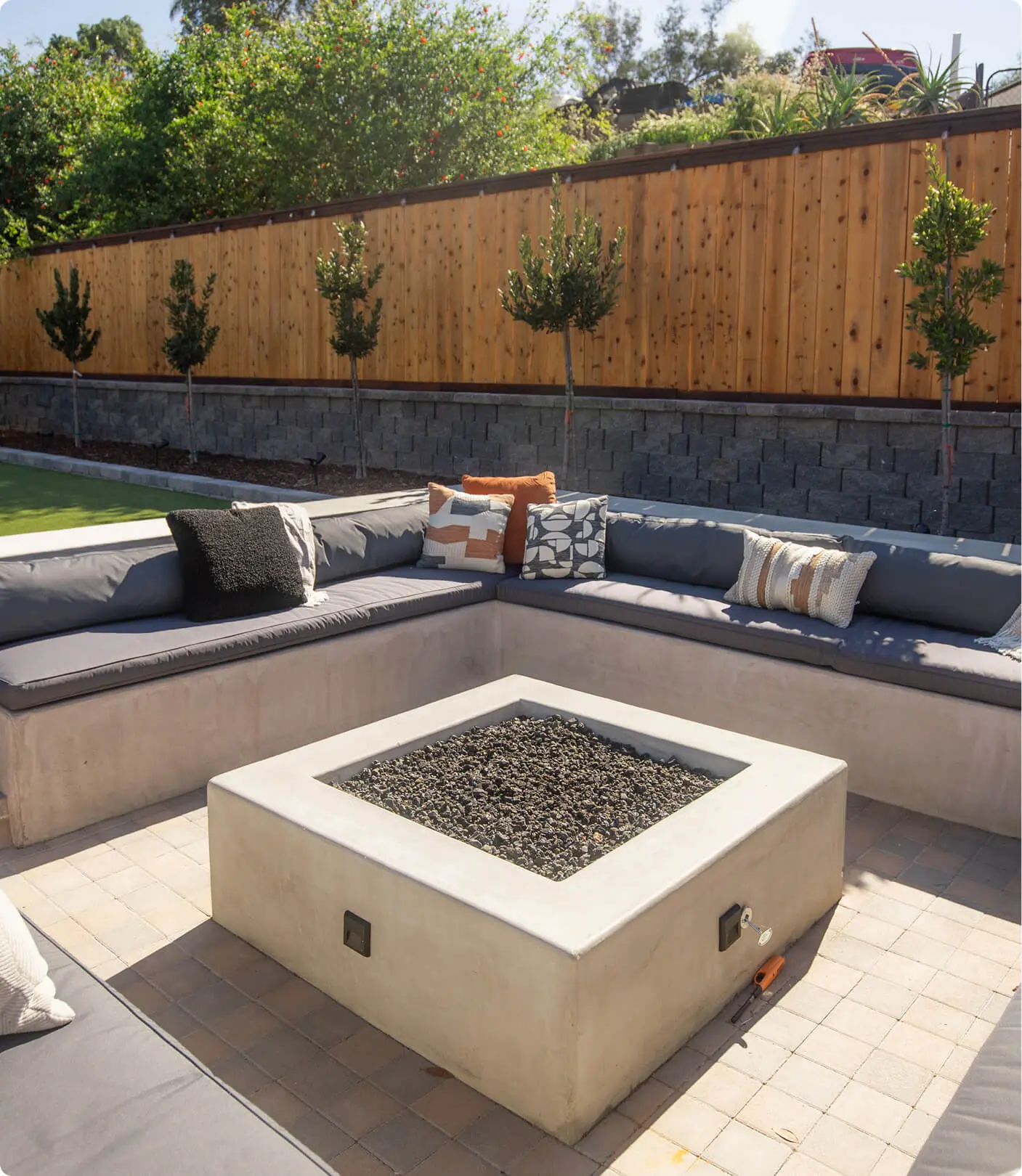 Sunken fire pit lounge by Sunshine Collective Studio featuring a large square concrete fire pit with lava rock, gray cushioned built-in bench, patterned throw pillows, and a row of topiary trees along a cedar fence.