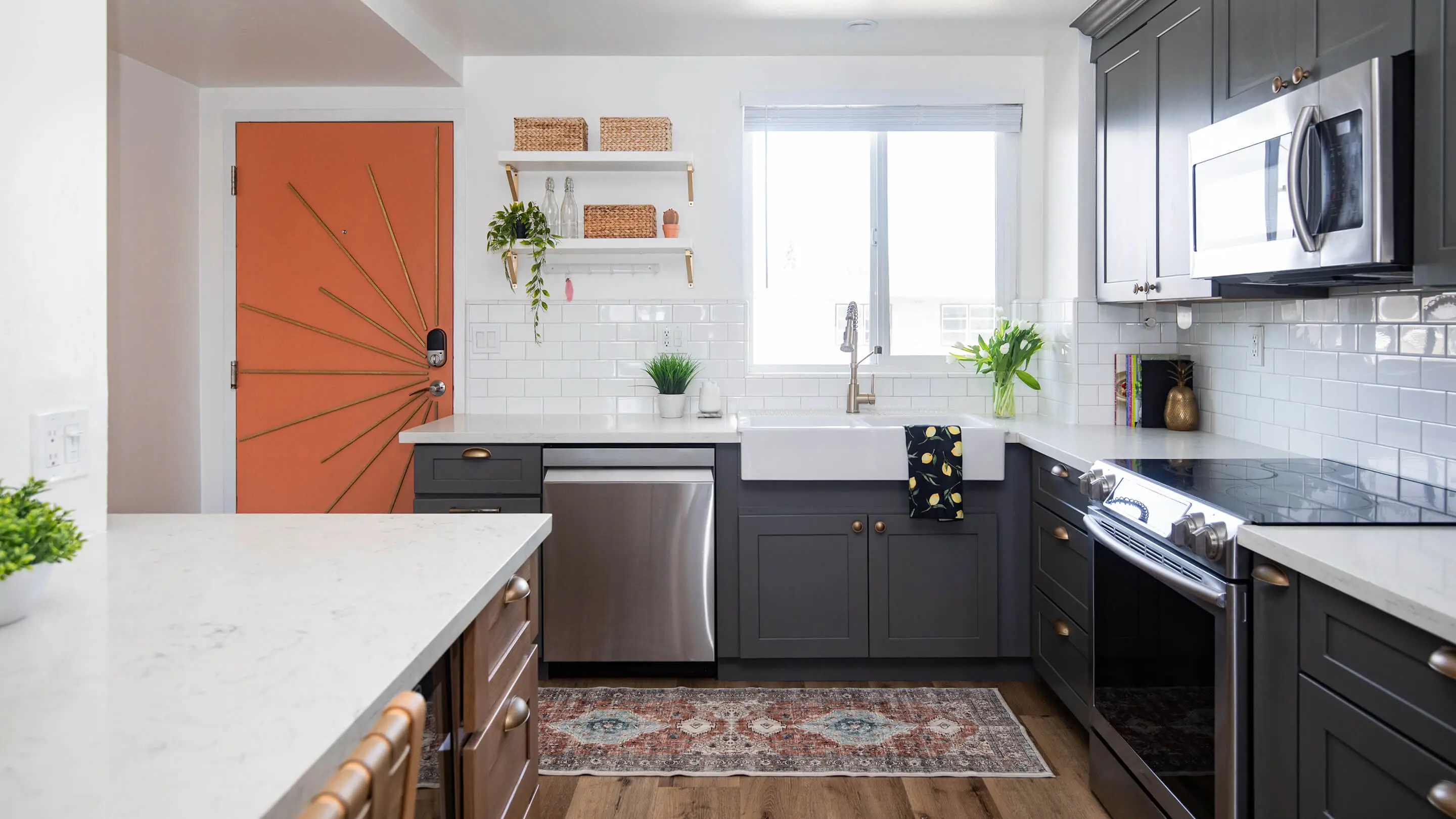 Renovated condo kitchen by Sunshine Collective Studio featuring gray shaker cabinets, brass hardware, farmhouse sink, marble countertops, white subway tile, open shelving, and a bold terracotta sunburst front door.