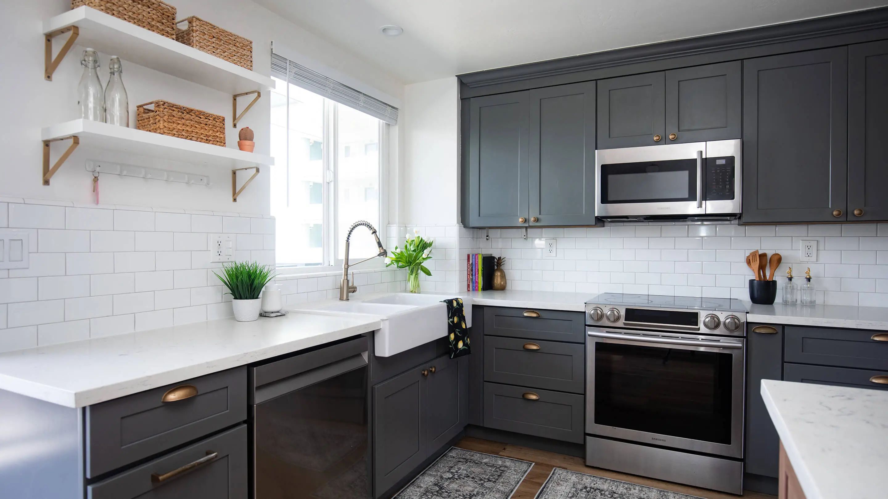 Kitchen by Sunshine Collective Studio with gray shaker cabinets, brass hardware, farmhouse sink, white subway tile, quartz countertops, open shelving with woven baskets, and stainless appliances.