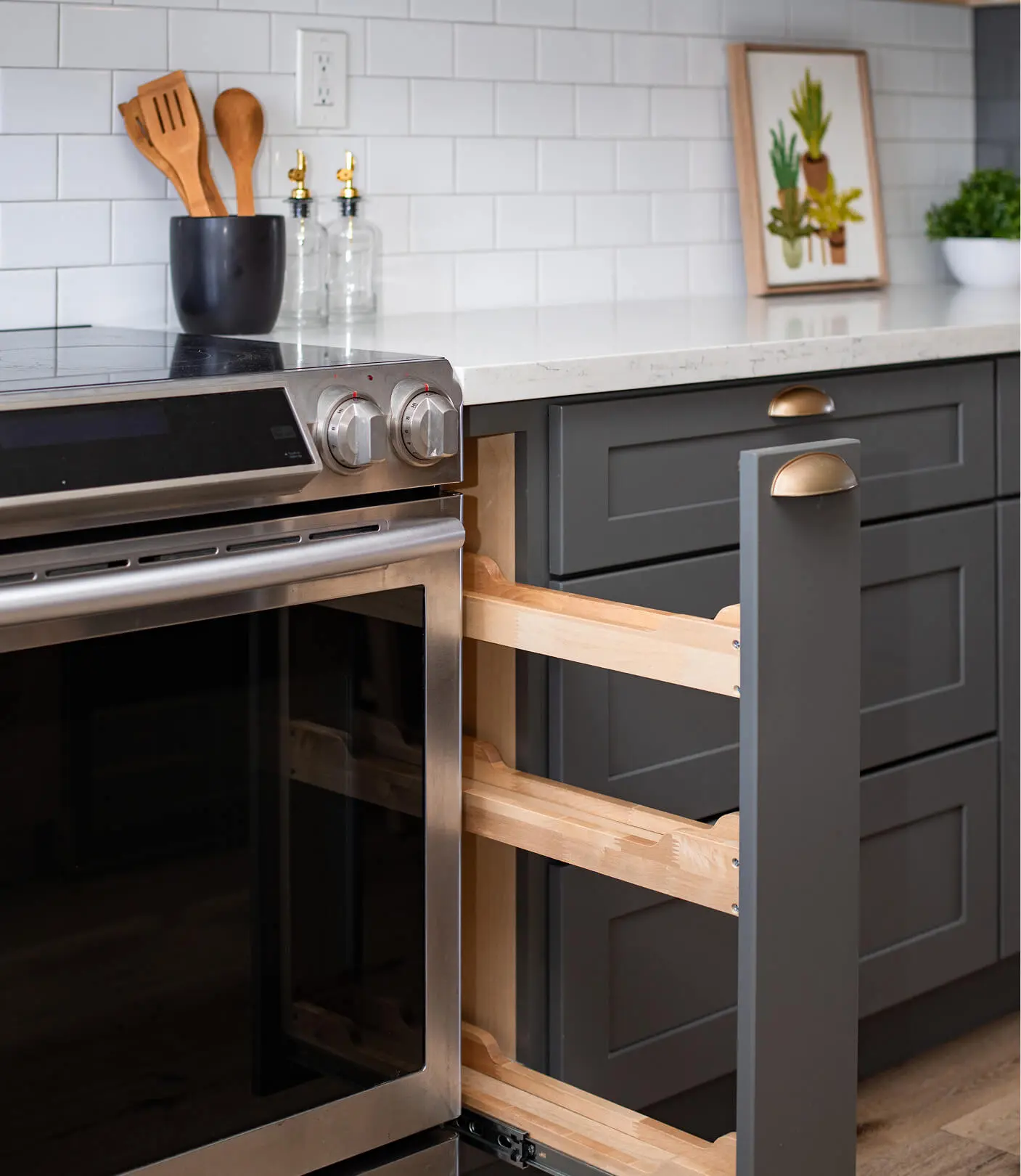 Kitchen storage detail by Sunshine Collective Studio showing a pull-out wood spice rack beside a stainless range, gray shaker cabinets with brass cup pulls, and white subway tile backsplash.