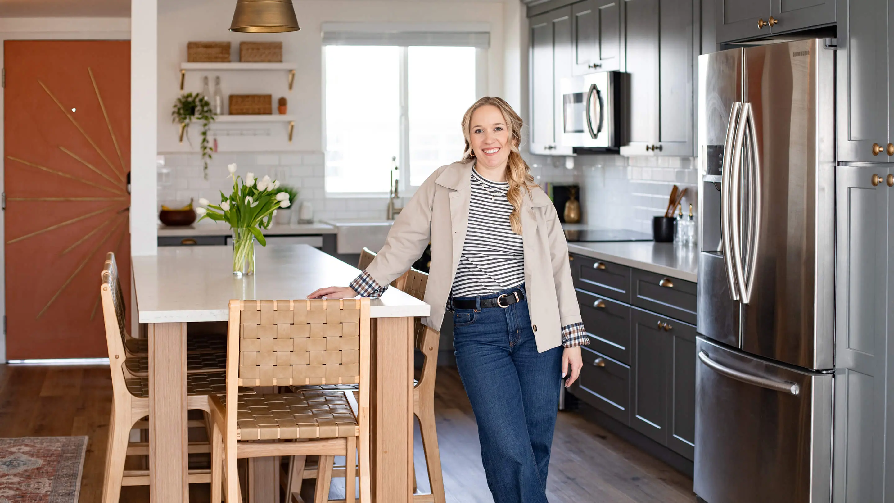 Founder and interior designer Kallie Morales of Sunshine Collective Studio stands at a marble kitchen island with woven leather bar stools, gray shaker cabinets, and a bold terracotta sunburst front door.