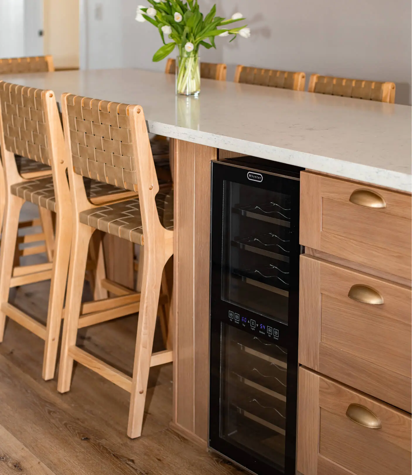 Kitchen island detail by Sunshine Collective Studio featuring a marble countertop, built-in dual-zone wine fridge, warm oak cabinetry with brass cup pulls, and woven leather bar stools.