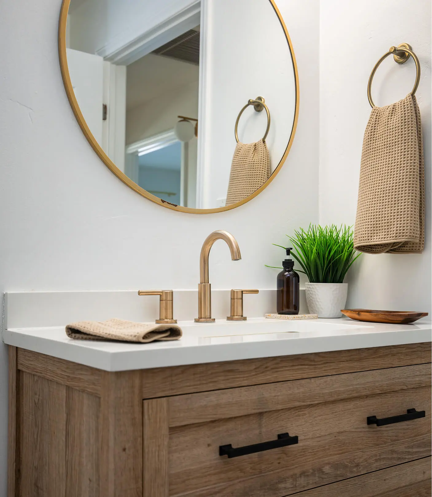 Bathroom vanity detail by Sunshine Collective Studio with a wood-tone cabinet, white quartz countertop, brass gooseneck faucet, round gold mirror, brass towel ring, and a small potted grass plant.