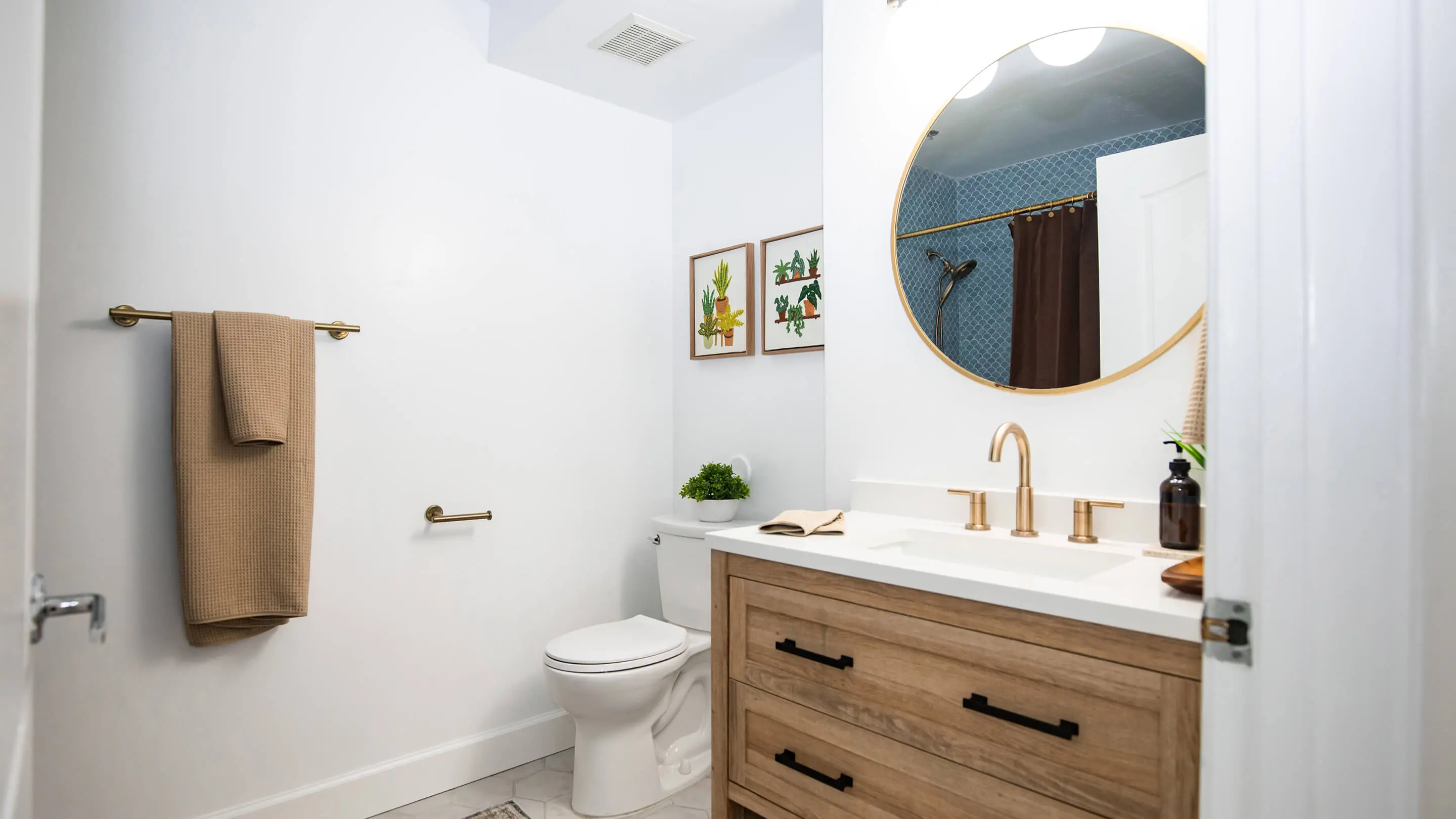 Bathroom by Sunshine Collective Studio with a wood-tone vanity, white quartz countertop, brass gooseneck faucet, round gold mirror, and a tub-shower with blue scallop tile and chocolate linen curtain.