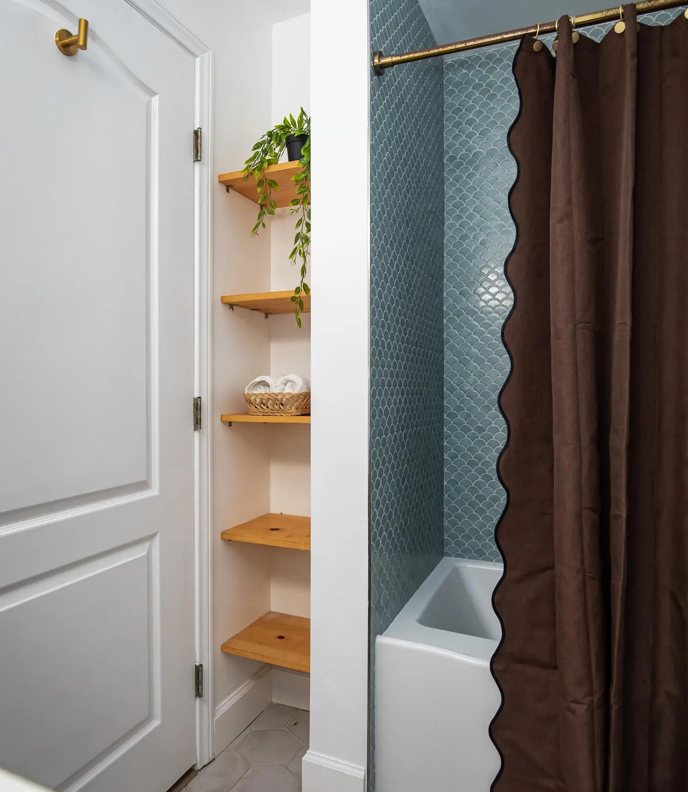 Bathroom by Sunshine Collective Studio with built-in wood corner shelving, a trailing plant, woven basket with rolled towels, and a tub-shower with blue scallop tile and scallop-edged chocolate curtain.