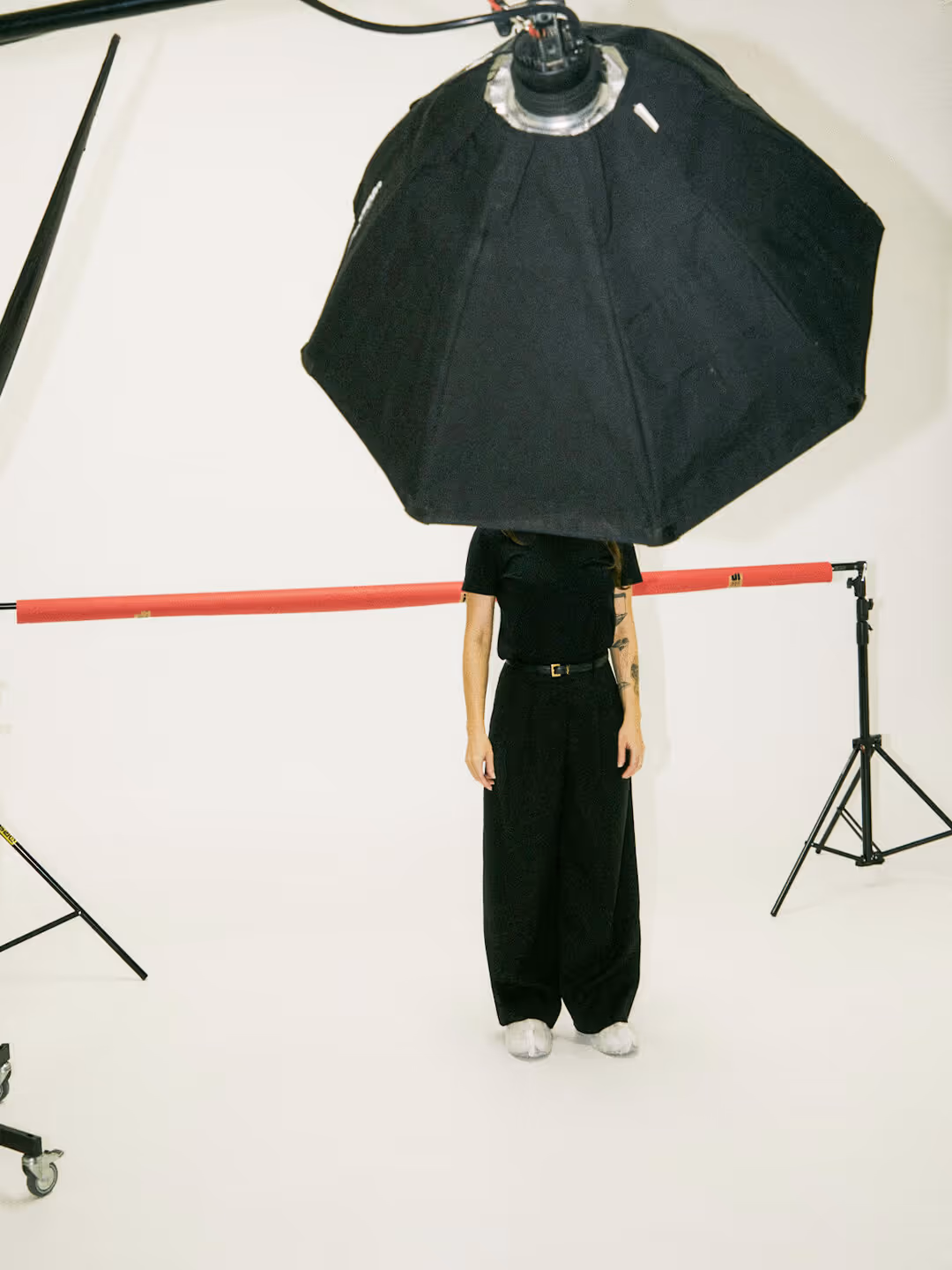 Person dressed in black standing behind a large black photography softbox that obscures their head, in a white studio with lighting equipment.