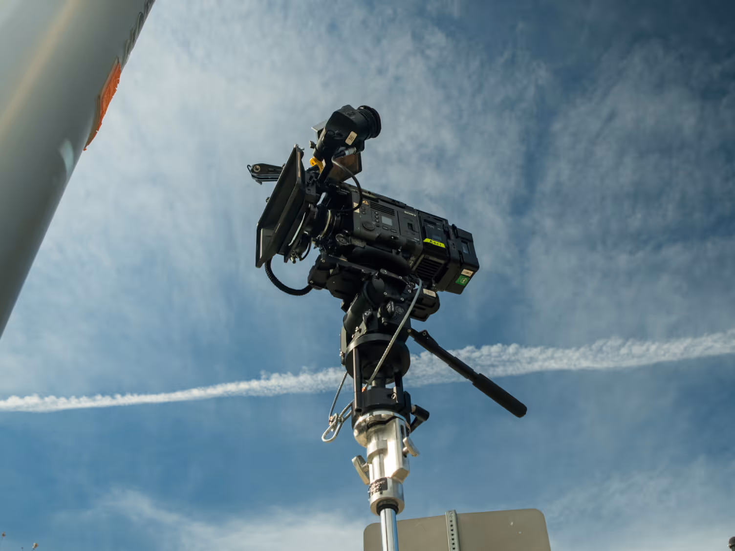 Professional video camera mounted on a tripod against a blue sky with wispy clouds and a jet contrail.