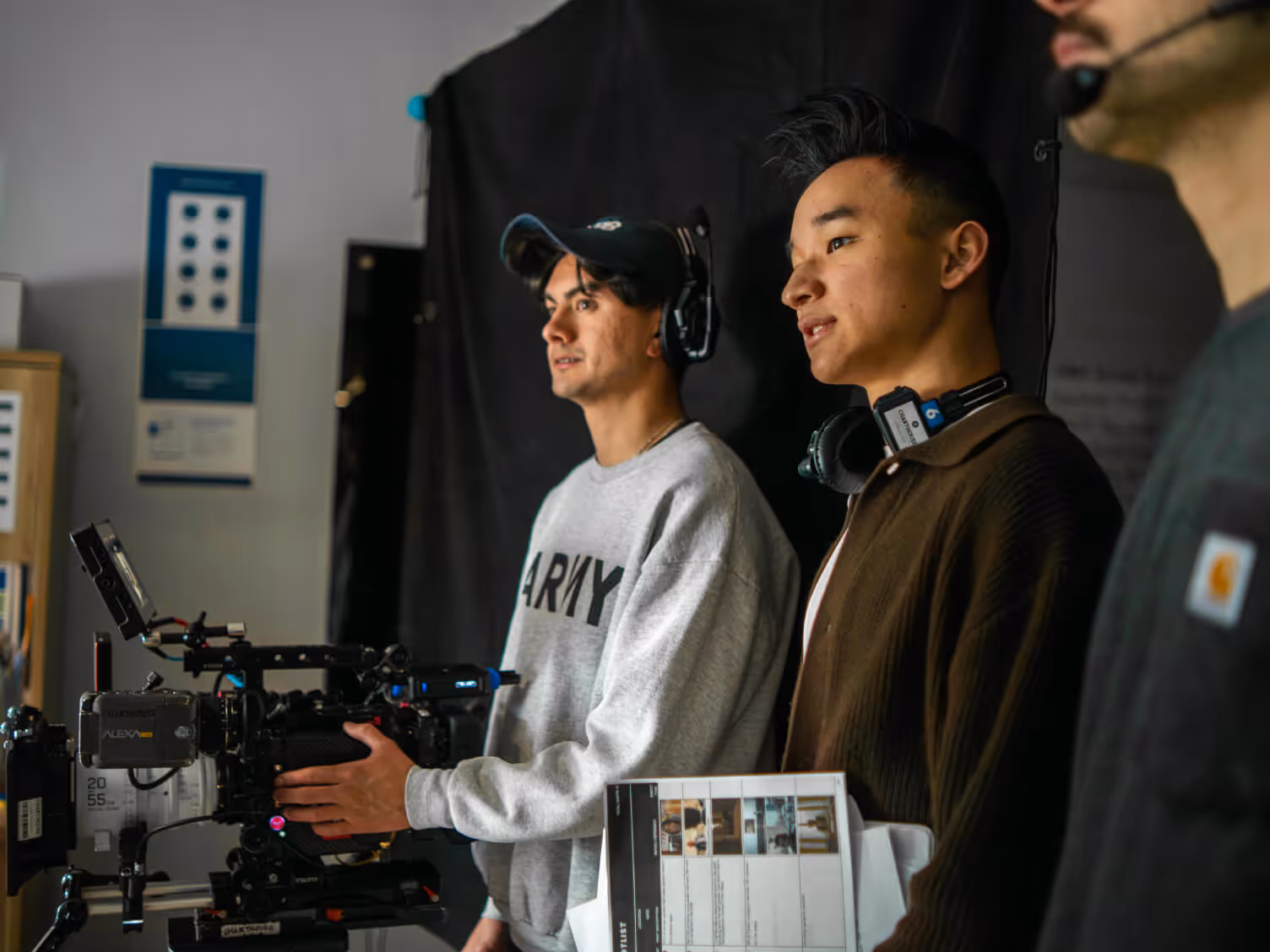Three young men on a film set with camera equipment, one holding a script, others wearing headphones and headsets.