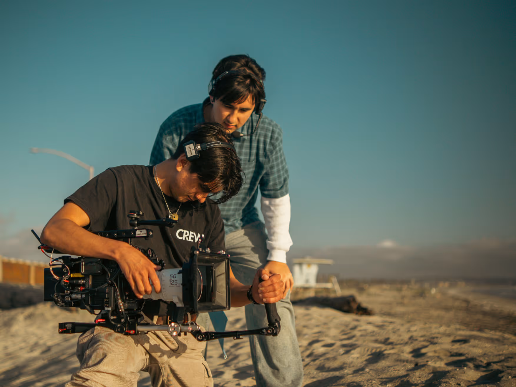 Two men on a sandy beach with one operating a professional video camera and the other watching closely.