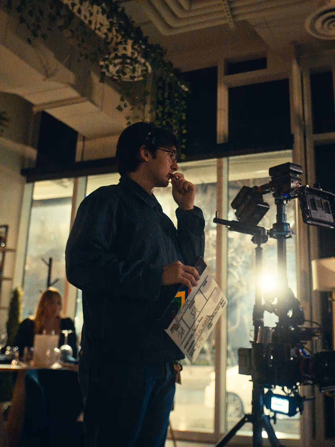 Person holding a clapperboard and wearing glasses on a film set with camera equipment indoors.