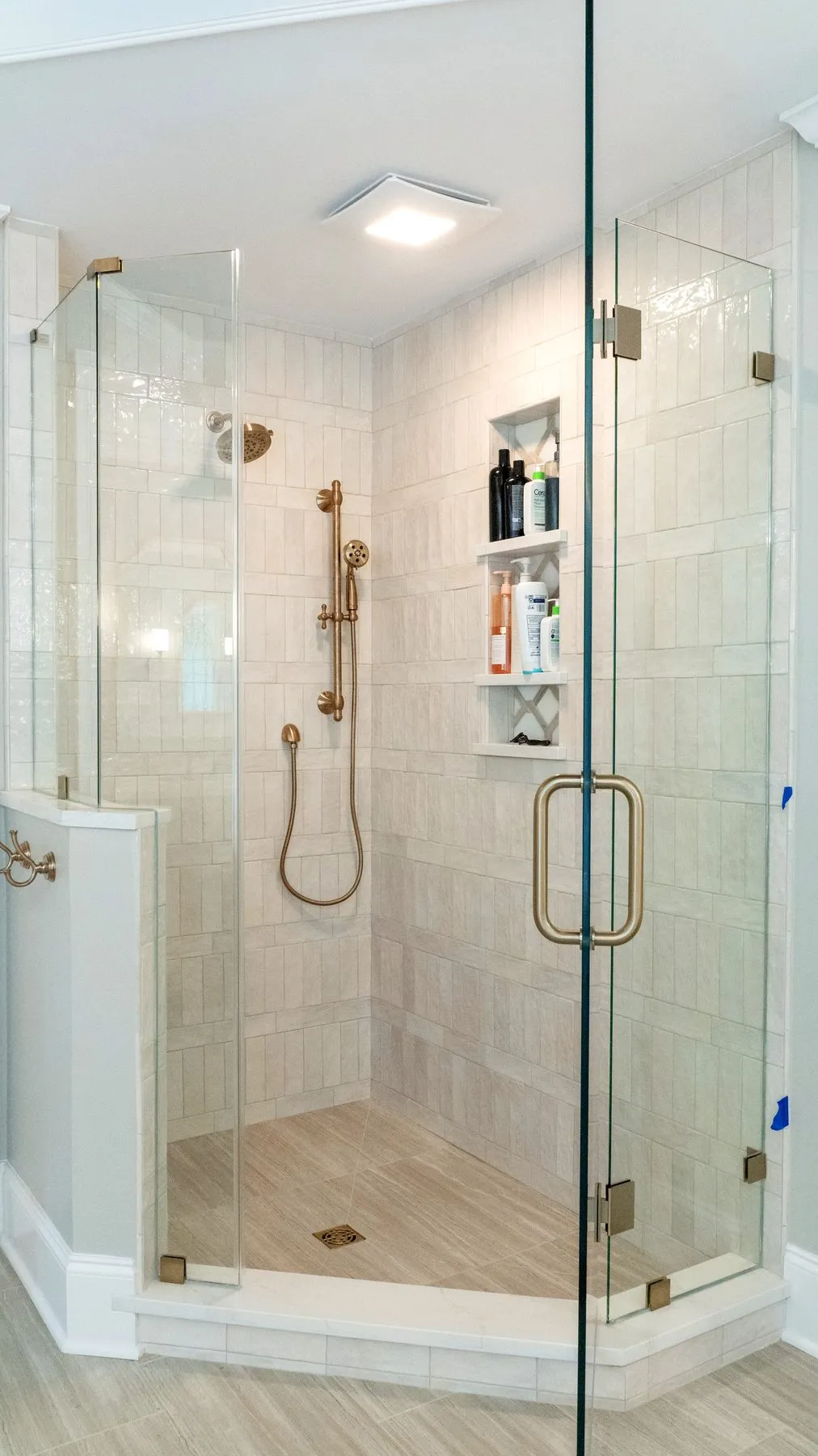 Walk-in shower tiled in travertine-look porcelain with brushed brass fixtures and a glass door