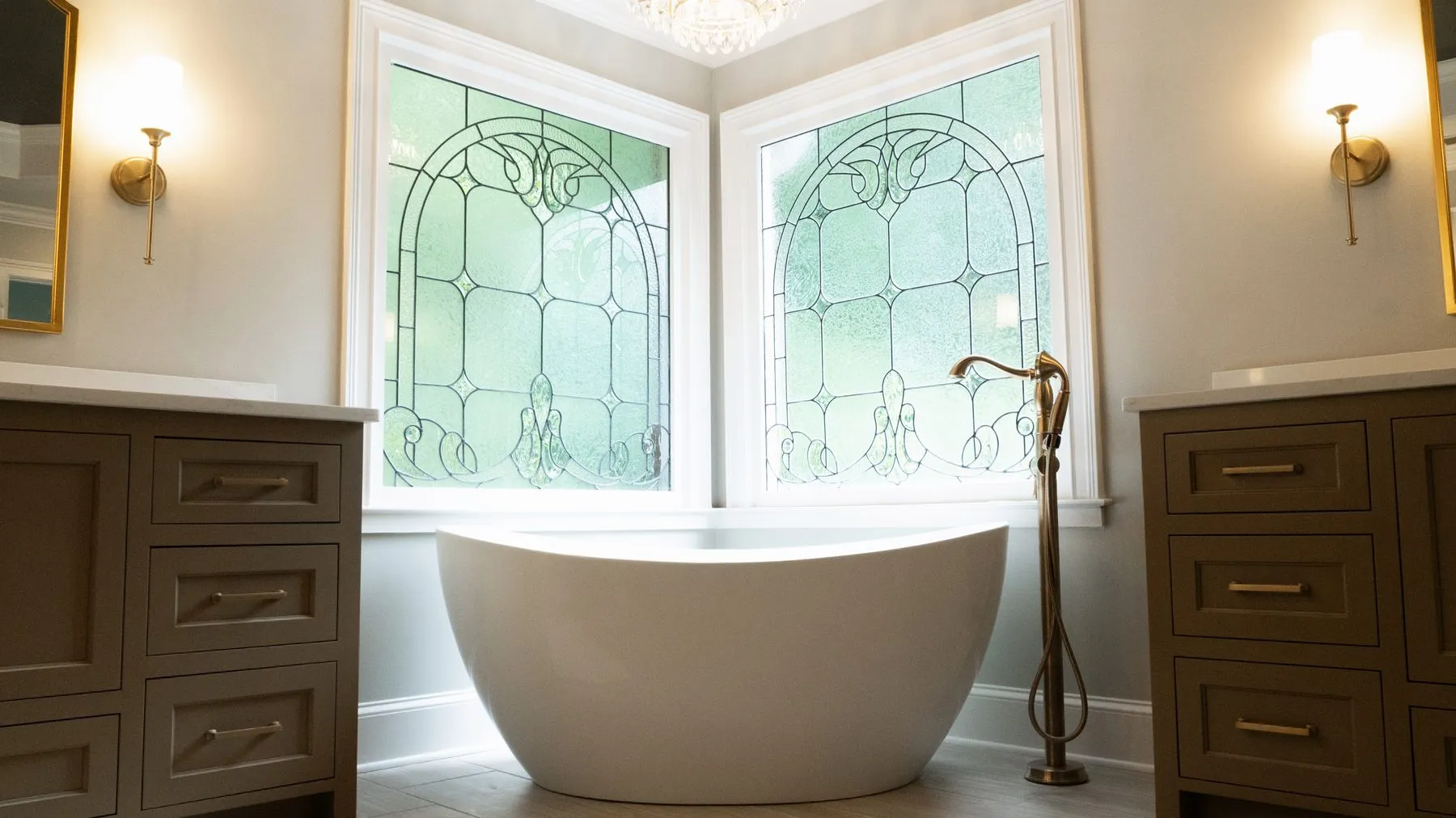 Freestanding tub set between two vanities beneath leaded glass windows, with brass fixtures