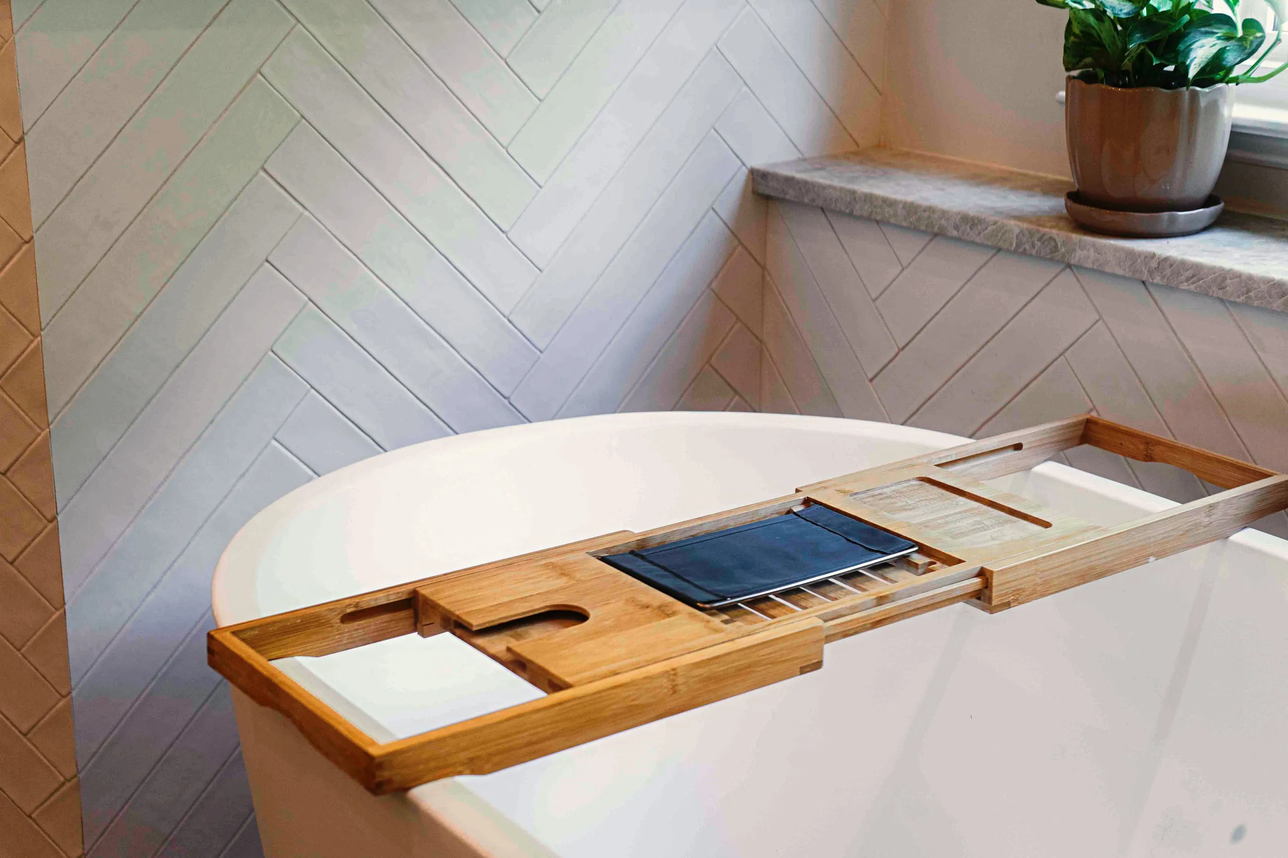 Freestanding tub against a herringbone tile wall with a wooden bath caddy