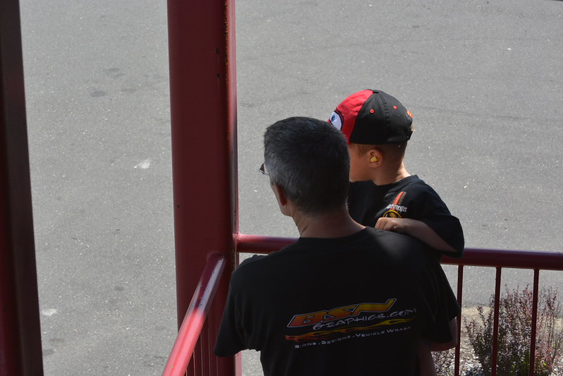 Man with gray hair holding a child wearing a red and black cap and yellow earplugs, standing near a red railing overlooking a gray pavement.