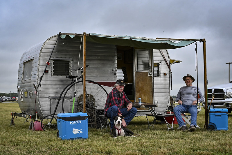 Two men sitting outside a vintage silver camper with an awning, with a dog sitting in front and blue coolers on the grass.