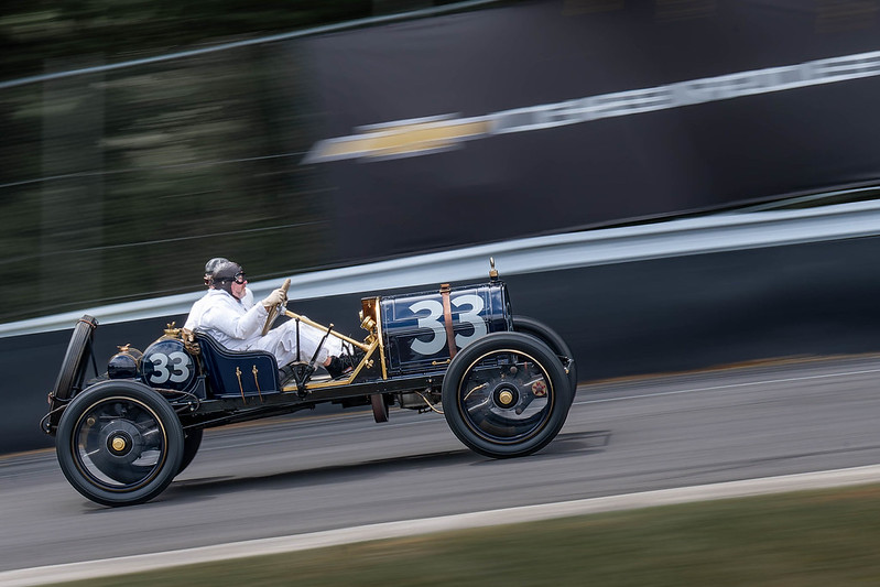 Vintage race car number 33 in motion on a racetrack with driver wearing white racing suit and helmet.