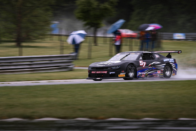 Black and blue race car number 57 speeding on a wet race track with spectators holding umbrellas in the background.
