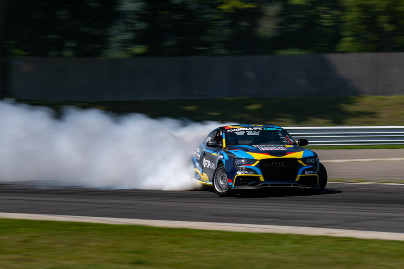 Blue and yellow racecar drifting on a track with smoke trailing behind its rear tires.