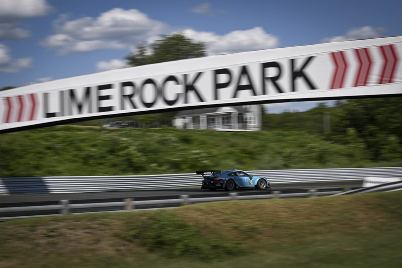 Blue race car speeding on Lime Rock Park race track under a bridge sign with greenery and blue sky in the background.