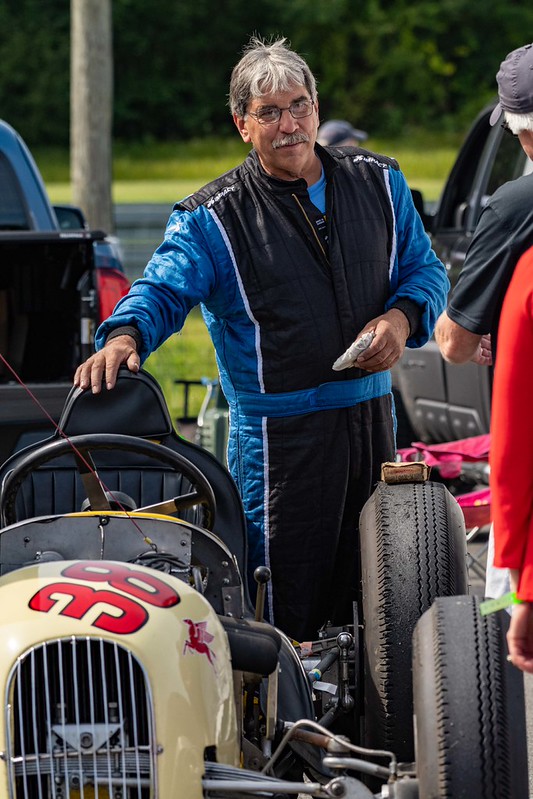 Man in blue and black racing suit standing next to a vintage race car with the number 38 on the hood.