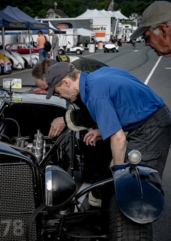 Two men closely inspecting the engine of a vintage black car at an outdoor event with tents and other cars in the background.