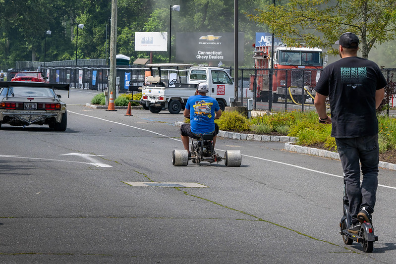 Man riding a small three-wheeled vehicle on a paved road with another man riding an electric scooter nearby.