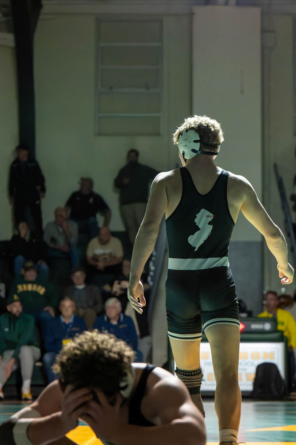Wrestler in black singlet and white headgear walking away on mat while opponent kneels with head in hands.