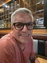 Smiling man with gray hair wearing black glasses and a salmon-colored T-shirt sitting in a restaurant booth with a mirrored wall behind him.