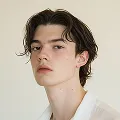Portrait of a young person with wavy brown hair and fair skin wearing a white shirt against a light background.