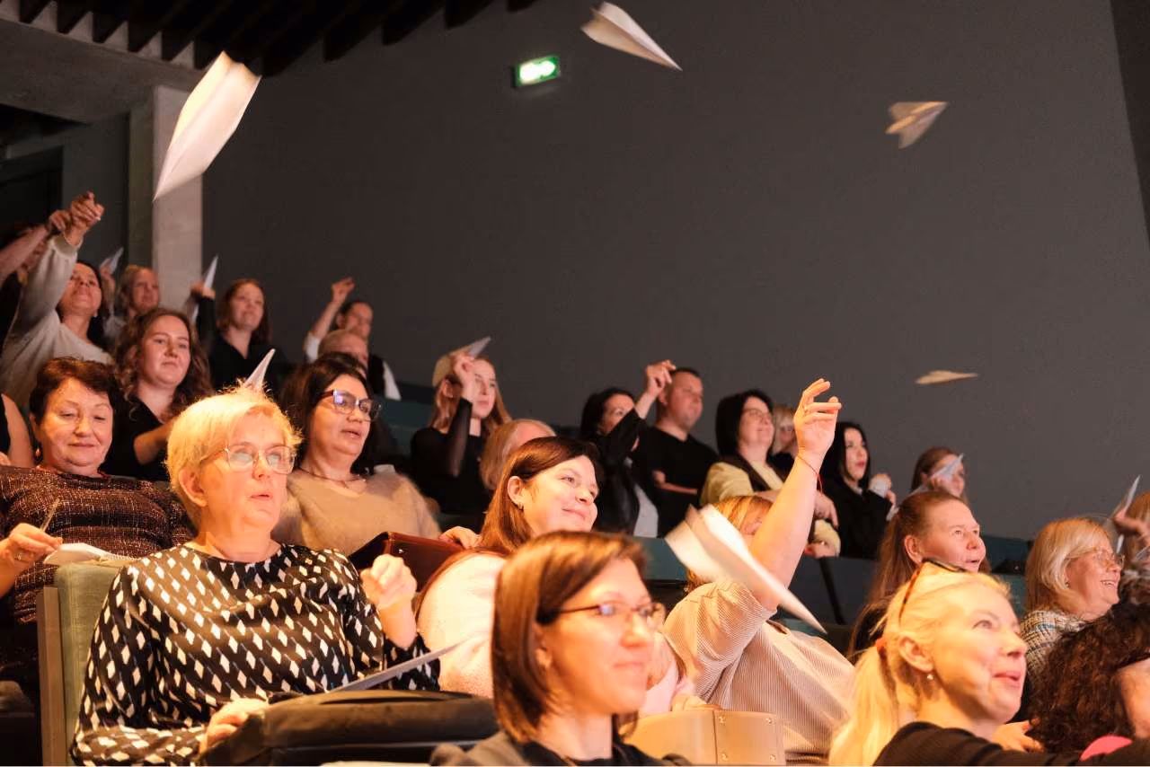 Auditorijā cilvēki smaida un laiž papīra lidmašīnas.