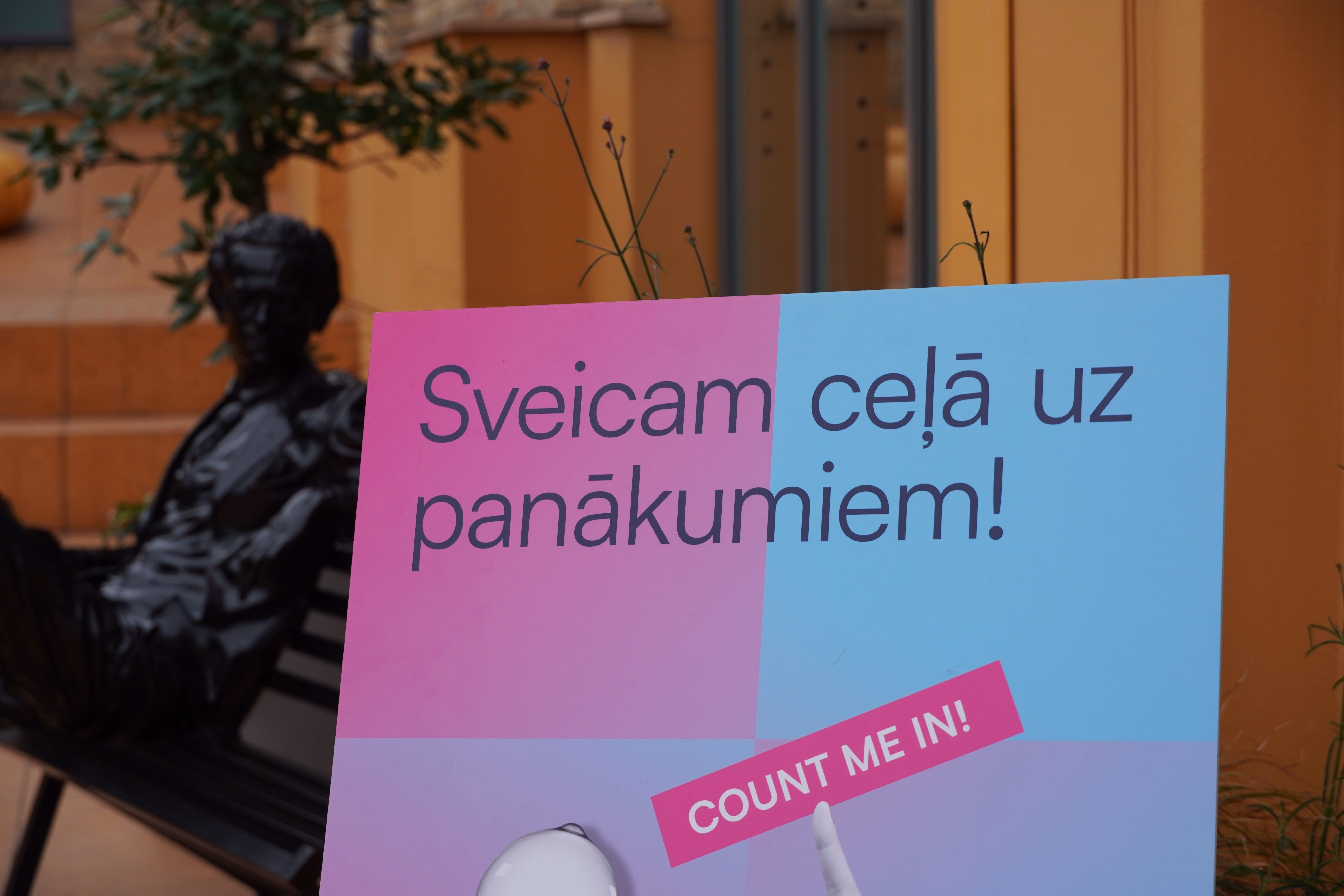 Sveiciena plakāts rozā un gaiši zilos toņos ar uzrakstu "Sveicam ceļā uz panākumiem!" un rozā lenti ar tekstu "COUNT ME IN!". Kreisajā pusē fonā redzama tumša vīrieša skulptūra, kas sēž uz soliņa.