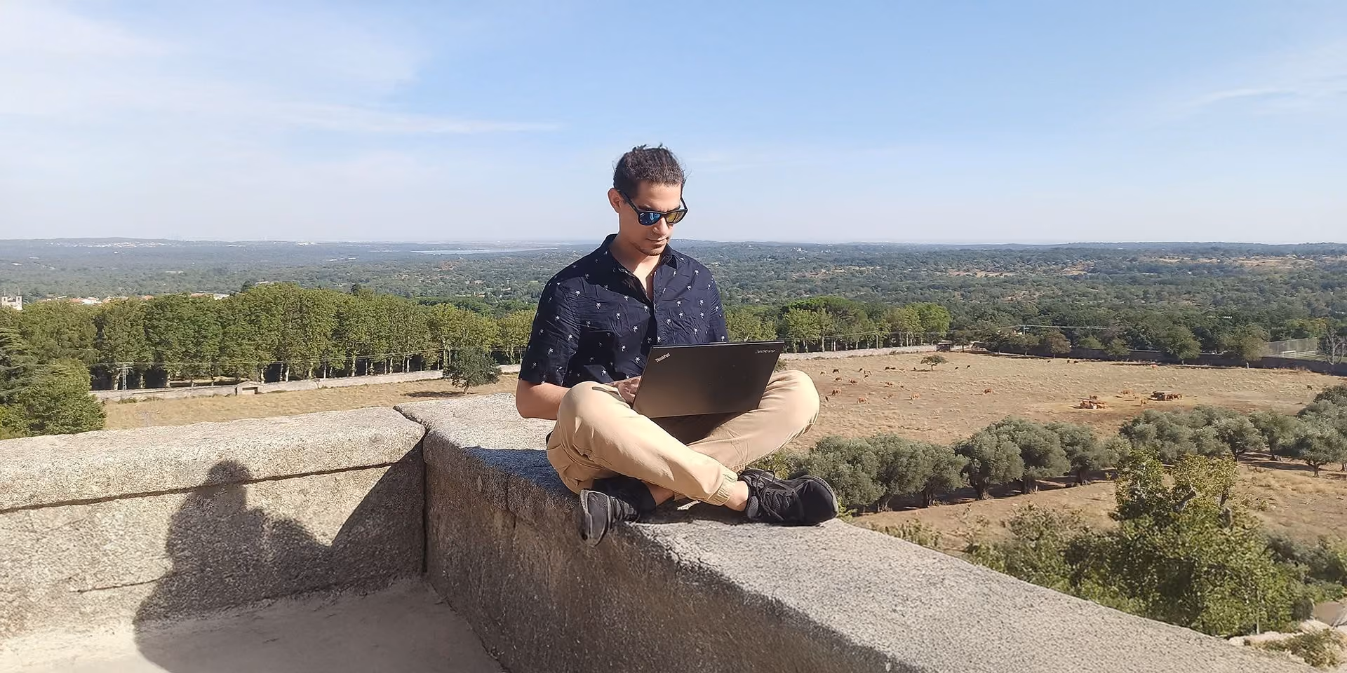 Cilvēks sēž uz akmens krasta laukuma ar portatīvo datoru, fonā plašs mežu un zemes ainavas skats.
